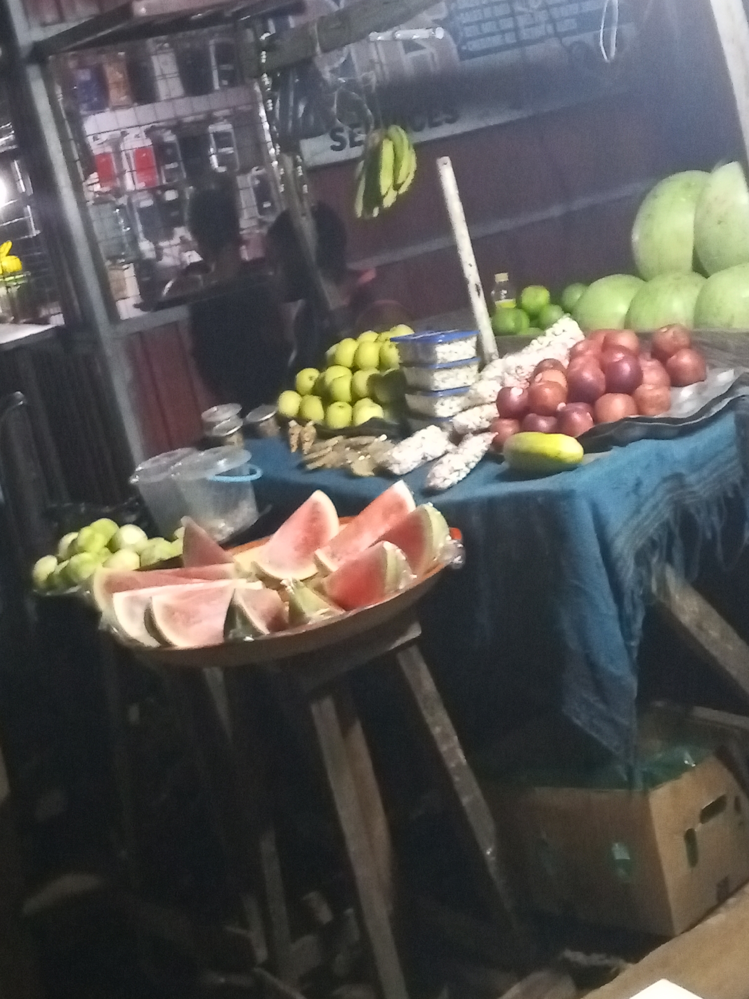 A street vendor's stall in Lokoja, Nigeria, displays a variety of fruits on a blue cloth-covered table. Watermelon slices are presented on a plate set on a wooden stool, while other fruits such as oranges, limes, and green gourds are arranged on the table. Packaged goods in stacked containers are also visible, along with a lone mango. Two people are standing near the stall. A storefront with phone cases and accessories behind the vendor suggests the street location is a commercial area.