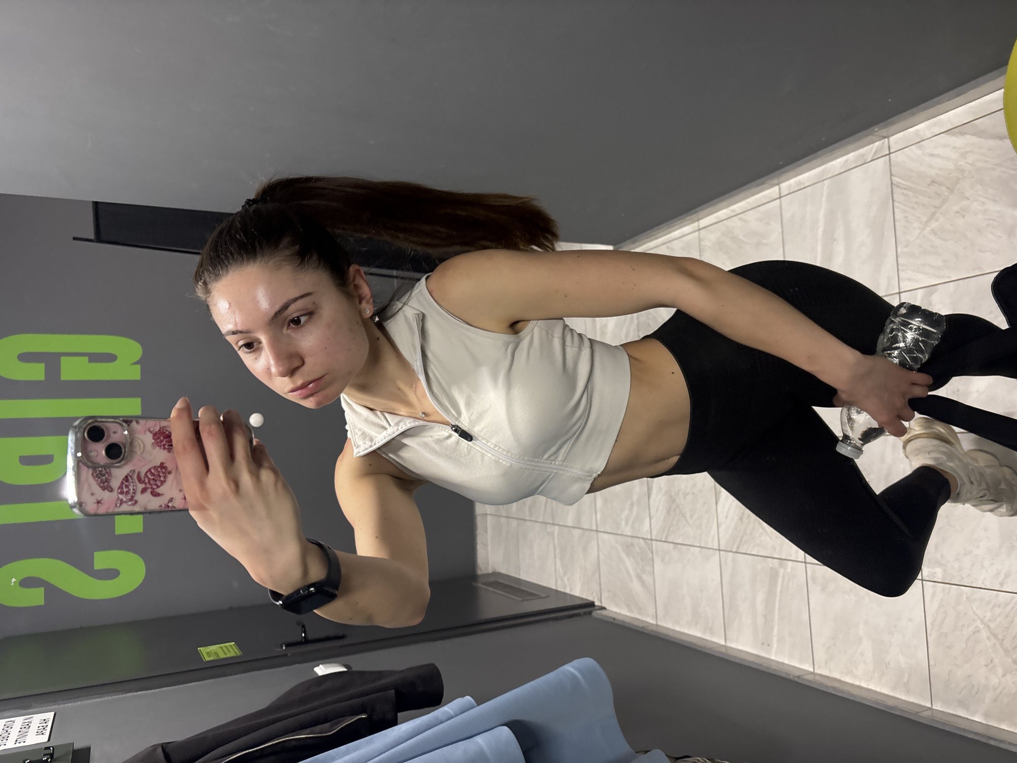 A woman is captured in a mirror selfie inside what appears to be a gym or fitness center. She has long, dark hair tied in a high ponytail. She is wearing light-colored athletic attire, including a sleeveless crop top with a zippered front and black leggings. A black smartwatch is visible on her left wrist.
In her right hand, she holds a smartphone with a pink, patterned case, angled towards the mirror to take the photo. Her left hand is holding a clear plastic water bottle. The woman's expression is neutral as she looks towards the phone screen.
The setting features grey walls and a light-colored tiled floor. On the grey wall, large green text is visible in reflection, partially obscured, appearing to read "GP" followed by other letters, possibly "D". Below this, on a darker panel, there is faint white text, also reflected, which is difficult to decipher fully but seems to spell out characters such as "NYKTRAINE" when viewed in reflection. In the foreground, at the bottom left, some dark and light blue fabrics, possibly clothing or towels, are visible. The lighting is bright and artificial, consistent with an indoor facility. The overall impression is that of someone pausing during a workout for a picture.