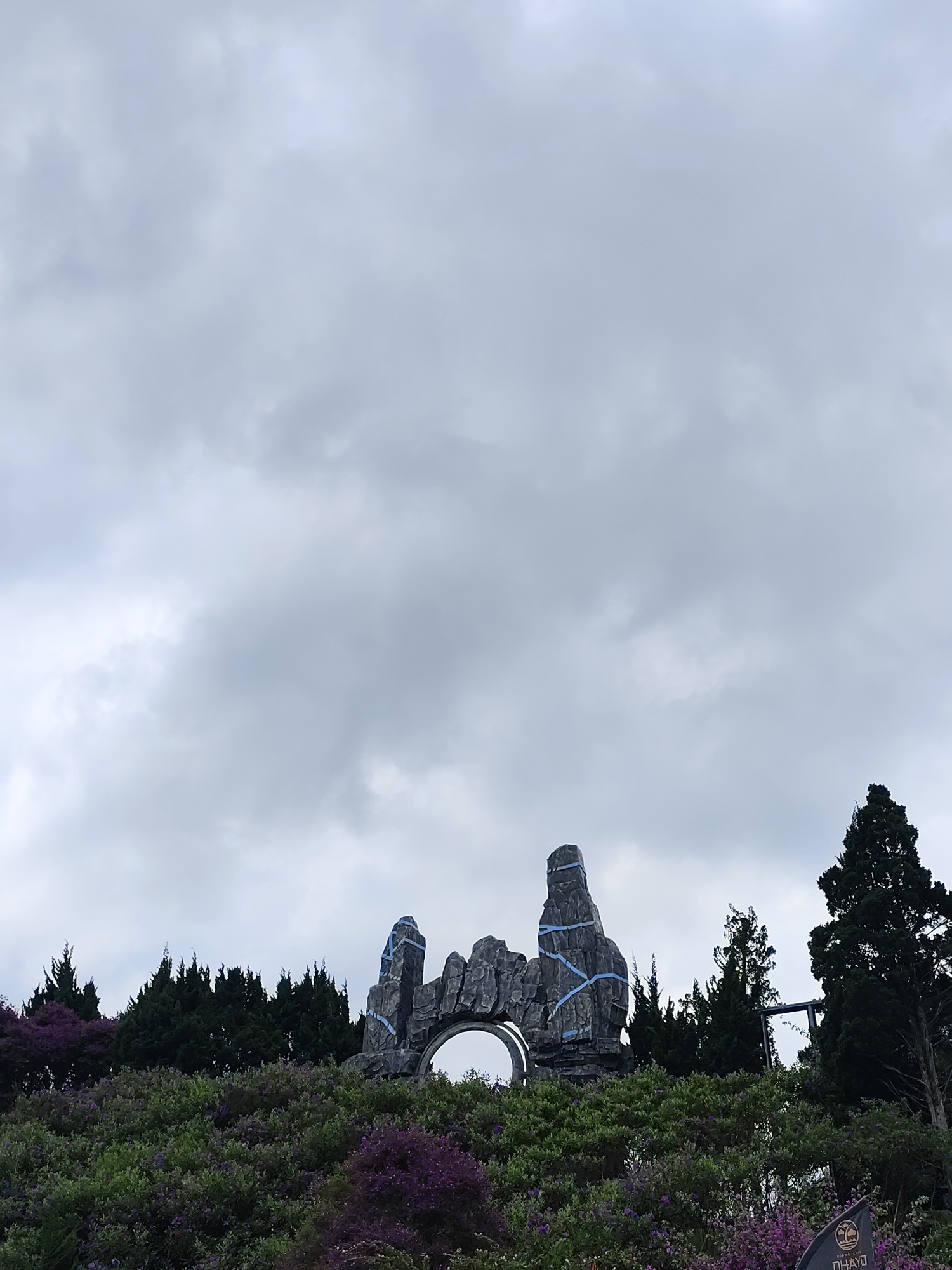The image presents an outdoor landscape under a heavily overcast, grey sky that dominates the upper two-thirds of the frame. In the midground, a prominent architectural feature stands out: a large, dark grey, rock-like structure resembling an archway or gate. This structure features two distinct vertical formations that suggest pillars or natural rock spires, with bright blue ribbons or tape wrapped around parts of it, adding a contrasting detail.
The foreground and midground are densely covered with lush vegetation. Vibrant green bushes are interspersed with clusters of purple flowering plants, possibly rhododendrons or azaleas, creating a rich texture. Behind the rock structure and along the horizon, a line of dark green evergreen trees, likely conifers, forms a natural backdrop.
In the bottom right corner, a dark, partially obscured sign is visible. The legible text includes "BOUNCY.CLUB" at the top, followed by "DA LAT", a circular tree logo, and another word below, possibly "PHAYO" or "NHAYO".
The scene appears still and tranquil, with no human activity visible. The soft, diffused light characteristic of the cloudy day contributes to a calm and contemplative atmosphere. The unique rock formation and abundant flora suggest a scenic park or garden in Da Lat, Vietnam, as indicated by the context.
