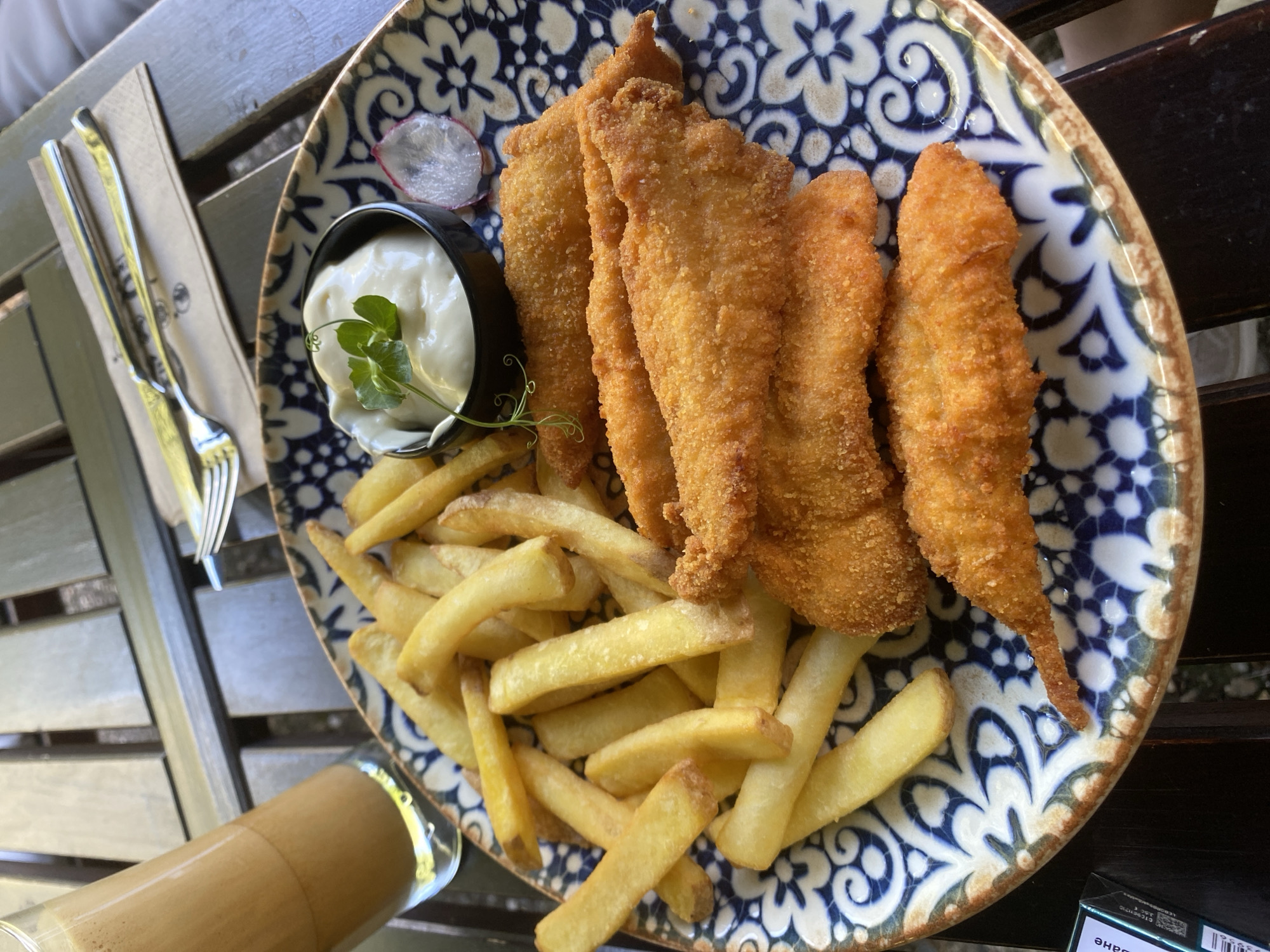 A close-up, overhead shot shows a plate of food on a wooden table. The plate is decorated with a blue and white patterned rim and contains a pile of golden french fries, several breaded fish fillets, a small dish of white sauce, and a thin slice of radish. A glass of iced coffee with a straw stands to the left of the plate. A fork and knife are placed on a napkin behind the plate. The background is out of focus but appears to be an outdoor setting with other wooden surfaces. The scene is set at the restaurant "Chill Out Bar & Dinner" in Varna, Bulgaria.