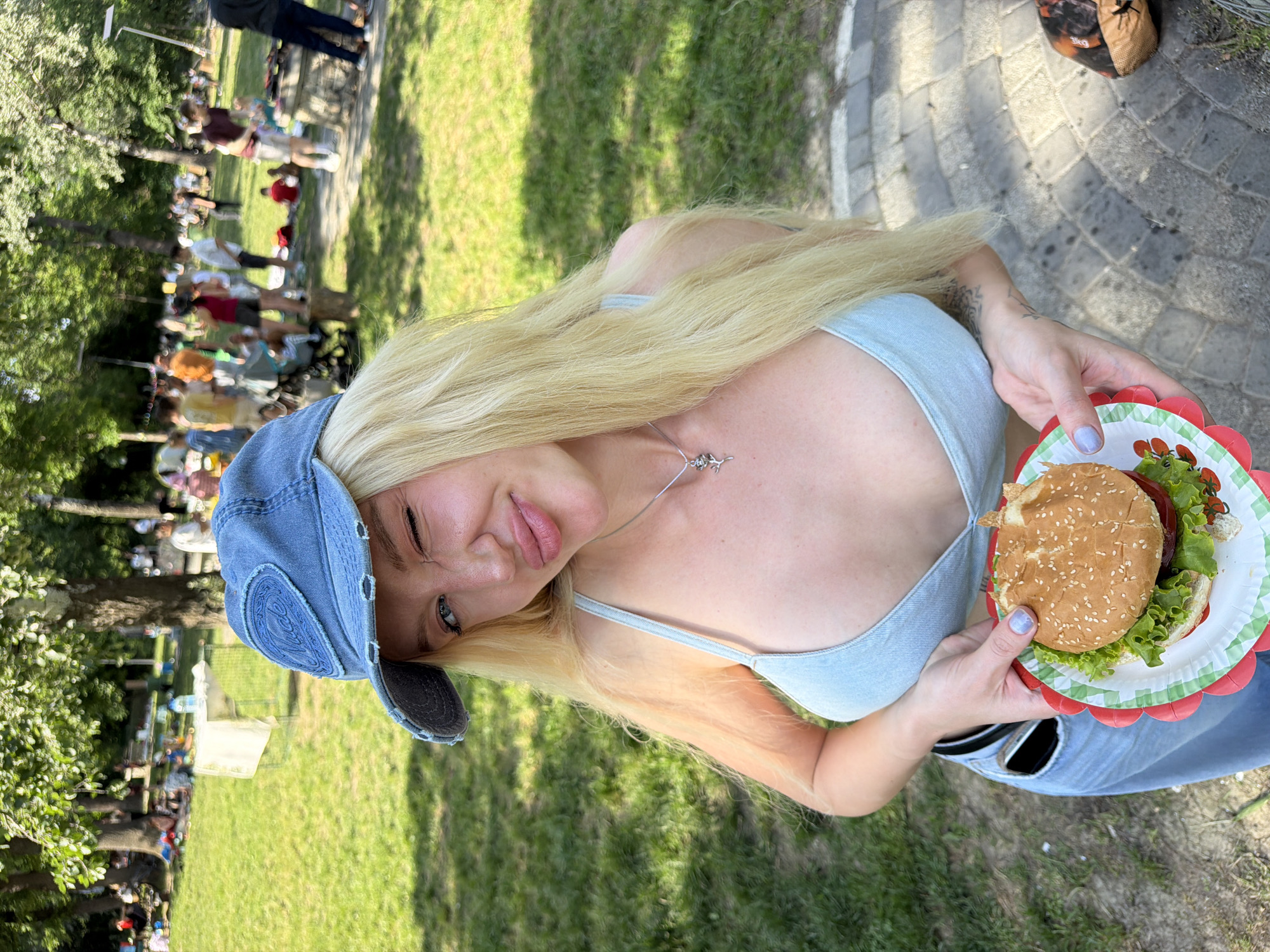 A fair-skinned woman with long blonde hair and a denim cap is holding a hamburger on a paper plate. She is in a grassy area with trees, and there are other people gathered in the background. The location appears to be outdoors, possibly a park. The background figures suggest an event or gathering, situated in what appears to be the Cukarica Urban Municipality, Serbia.