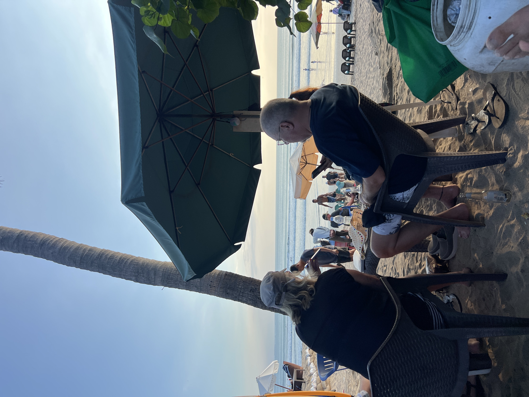 This image depicts a beachfront scene in Kuta, Indonesia, during what appears to be late afternoon or early evening. In the foreground, two individuals are seated at a table on the sandy beach, facing away from the camera towards the ocean. The person on the left, wearing a dark t-shirt and a baseball cap, has blonde hair. The person on the right, wearing glasses and a dark t-shirt, is looking down at a mobile phone. Behind them, other beachgoers can be seen in the middle ground, some walking near the water's edge, and others gathered around tables. A large, dark green umbrella shades the foreground seating area. A prominent palm tree trunk is visible to the left of the frame, and the ocean stretches out to the horizon, with gentle waves. Beach chairs are set up further down the beach. The overall atmosphere suggests a relaxed, social setting at a popular tourist destination.