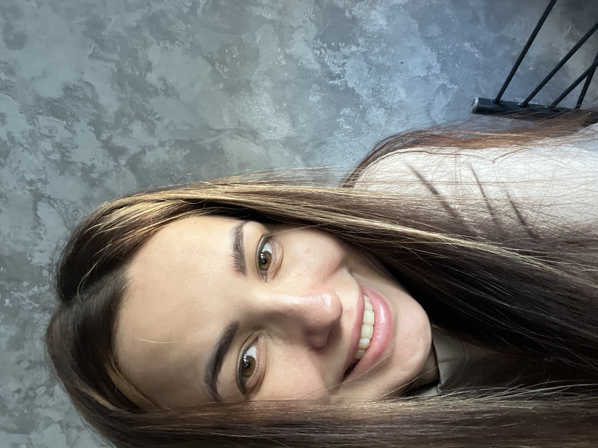 A woman with brown hair and eyes smiles towards the camera. She is wearing a light-colored shirt. The background is a mottled gray wall with a black metal object visible in the upper right corner. The location is unknown.
