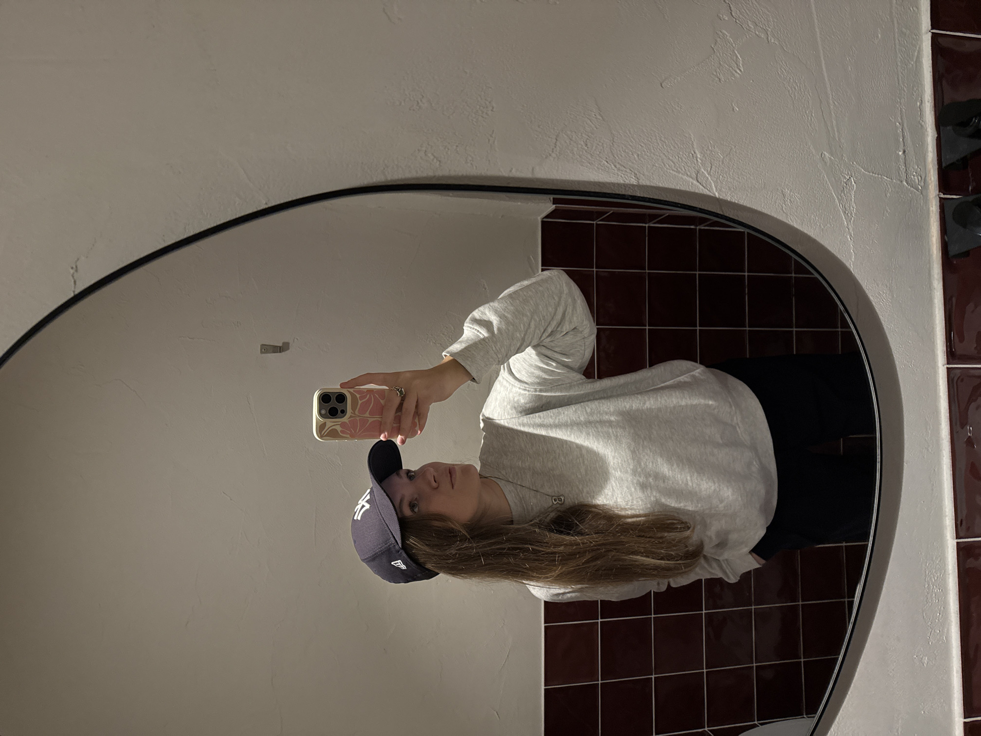The image depicts a young adult female taking a mirror selfie in an interior space in Belgrade, Serbia. The specific location within the city is not identifiable from visual cues. The subject, seen from the chest up, wears a grey long-sleeved sweatshirt, black trousers, and a navy blue New York Yankees baseball cap. Her long, light brown hair is visible beneath the cap. She holds a smartphone with a pink floral-patterned case in her right hand, angled to capture her reflection. A silver necklace with an '8' or 'B' shaped pendant is worn. Her left hand is partially visible, with fingers inside a pocket. The background consists of a large, irregularly shaped mirror with a thin black frame. The wall visible in the reflection has a lower section clad in dark red square tiles with white grout, surmounted by an off-white, textured plaster or stucco wall. A single metallic hook is affixed to the textured wall to the left of the reflection.