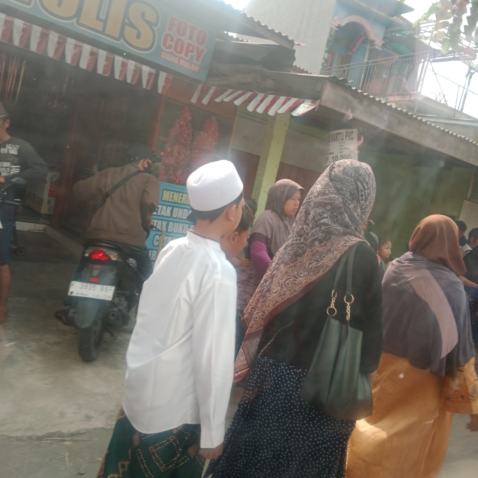 The image captures a street scene in Jekerto, Indonesia, likely a commercial area given the signage. In the foreground, a young boy wearing a white peci and a long-sleeved white garment is walking to the left. He is followed by another person, partially visible, also in traditional clothing. To the right of the boy, a woman in a dark hijab with a patterned scarf draped over it is walking away from the camera, carrying a dark green bag. Further to the right, another woman is seen from behind, wearing a tan hijab and an orange garment.
In the background, a man in a brown jacket and a dark cap is on a motorcycle, facing away from the camera. The motorcycle has a license plate that reads "K 3935 BBF". Above and behind the people, there are buildings with awnings and signs. One sign, partially obscured, appears to say "FOTO COPY" and "SIANG MALAM." Another sign reads "KARTU PVC" and "Rp. 10.000". The environment appears to be outdoors, possibly on a street or sidewalk, with a concrete ground surface. The lighting suggests it is daytime, with some harsh sunlight creating lens flare.