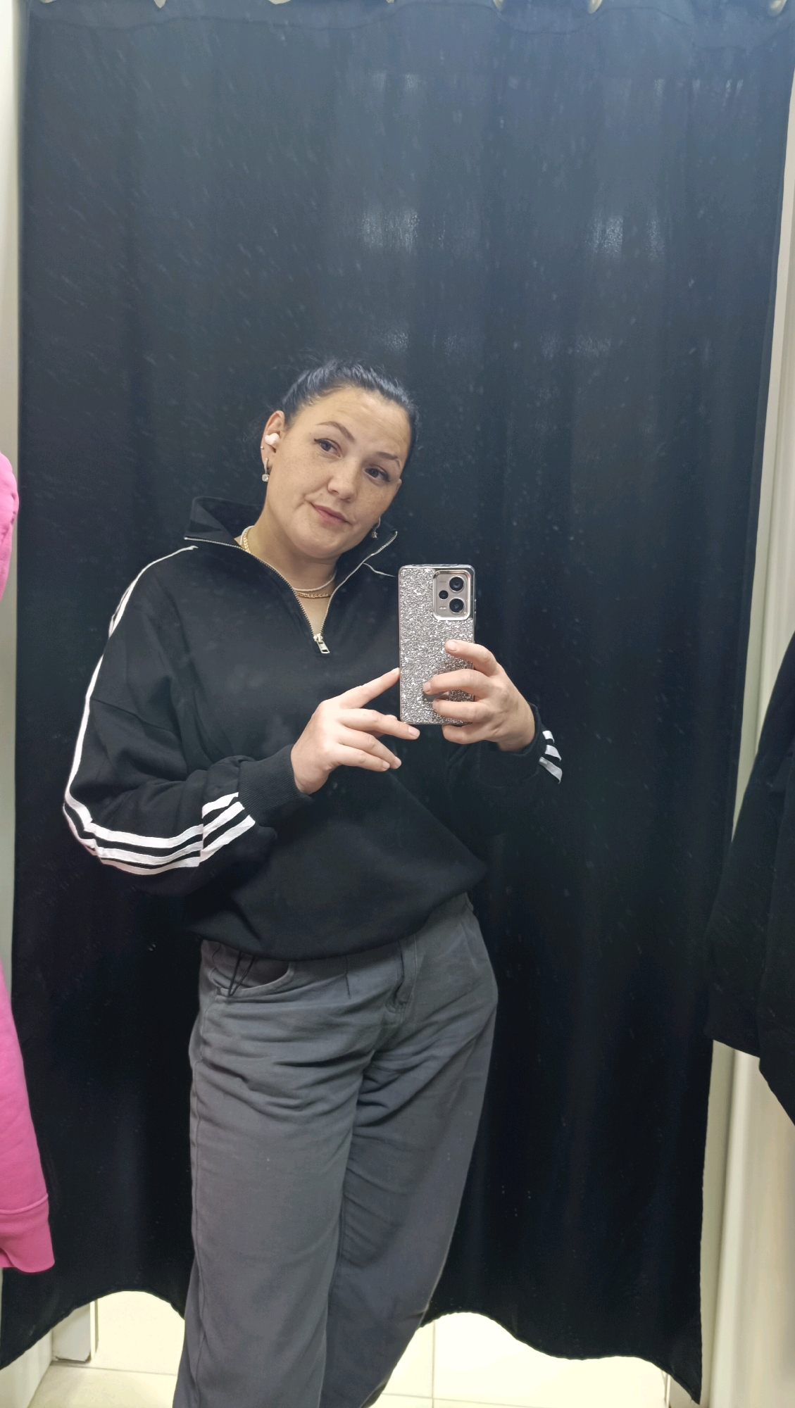 A woman is taking a selfie in a fitting room. She is wearing a black zip-up sweatshirt with white stripes on the sleeves and grey pants. She has dark hair pulled back and is wearing earrings and a necklace. She is holding a sparkly phone in her right hand. The background is a black curtain. The overall impression is of a casual, everyday scene.