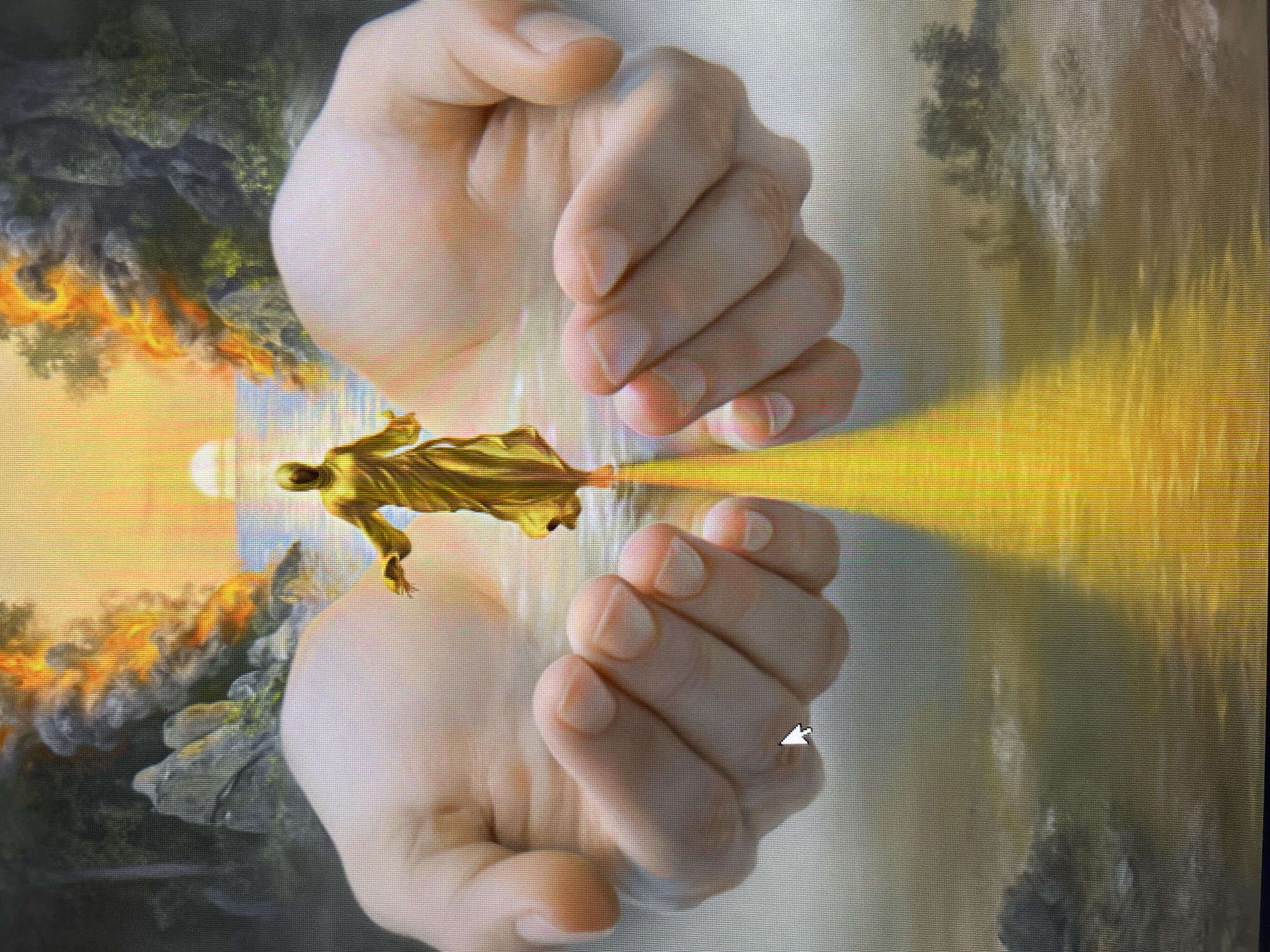A golden figure, resembling Jesus Christ, stands with outstretched arms in the center of the image, emerging from a shimmering yellow light. The figure is positioned between two large, cupped human hands that appear to cradle it. The background depicts a surreal landscape with rocky formations, water, and a fiery sky with a setting sun. The scene is artfully composed, creating a spiritual and ethereal atmosphere, without a specific city or location identified.