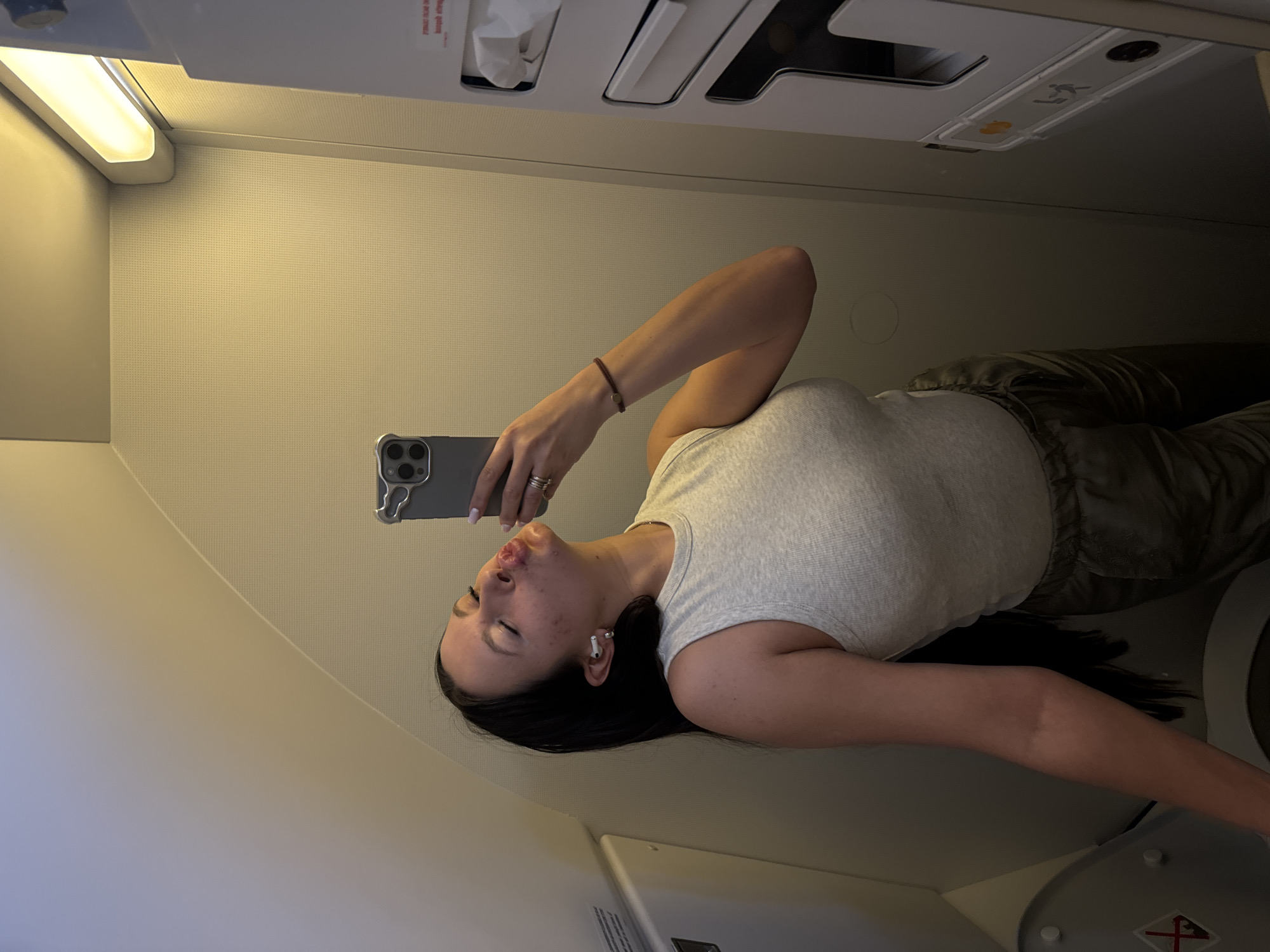 A woman is taking a selfie in what appears to be the cabin of an airplane. She's wearing a light-colored tank top and dark pants. She has dark hair and is wearing an AirPod in her left ear. She's holding a smartphone with a visible camera setup. The background includes the interior of the plane, featuring overhead compartments, reading lights, and other airplane fixtures. The location is not in Moscow, Russia, but in an airplane cabin.