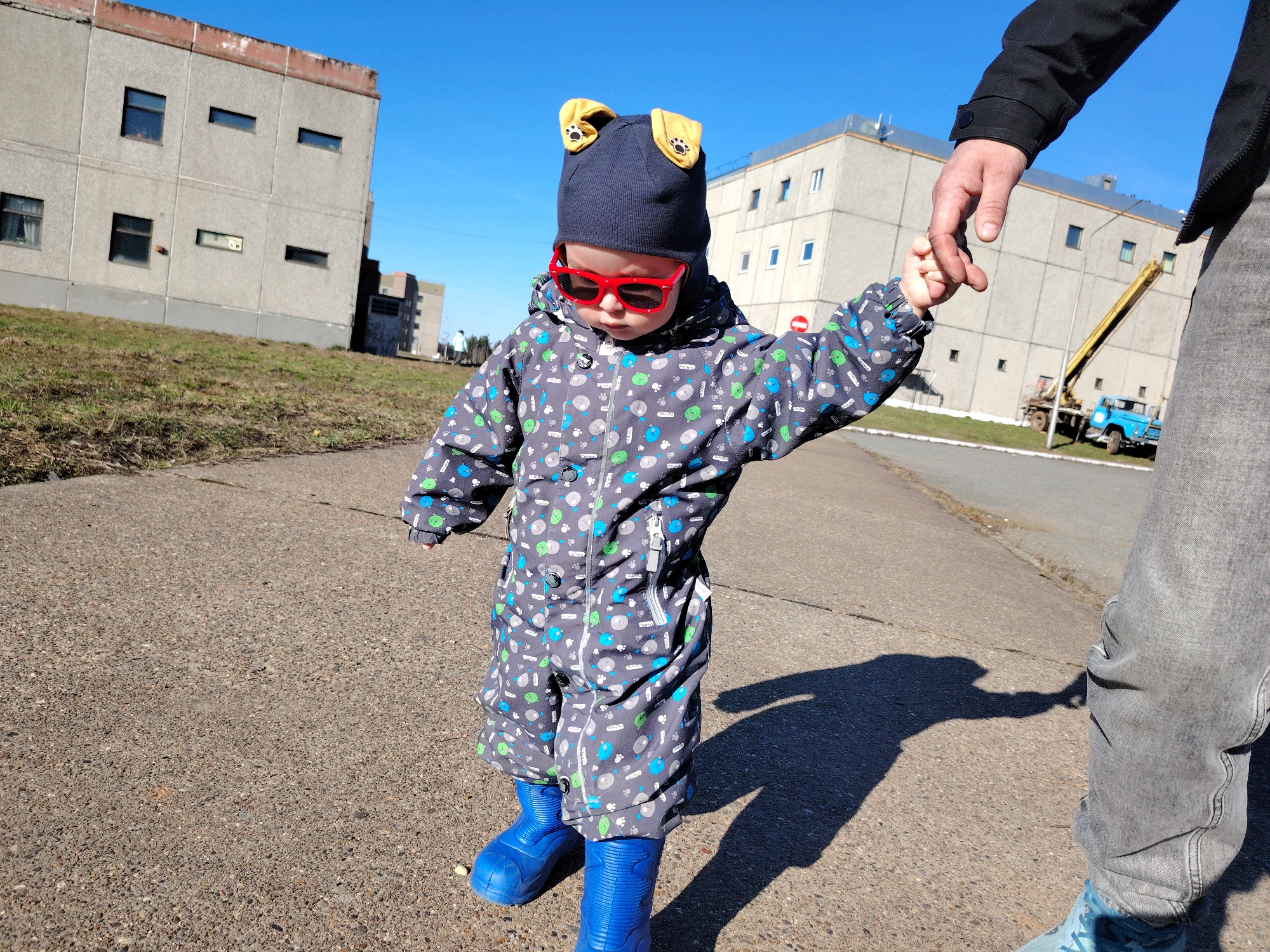 A small child, likely a toddler, is captured walking outdoors on a bright, sunny day, holding the hand of an adult. The child is dressed in a dark grey full-body jumpsuit adorned with blue, green, and light grey patterned circles and paw prints. They wear a dark blue beanie with prominent yellow dog ears featuring paw prints, bright red rectangular sunglasses, and chunky blue rubber boots. Only the adult's arm in a black jacket, hand, and lower leg in grey jeans are visible, indicating they are guiding the child.
They are walking on a paved surface, possibly asphalt or concrete, with patches of dry grass alongside. Strong shadows cast by the subjects suggest it is mid-day with clear, sunny weather and a vivid blue sky. In the background, several large, functional concrete buildings with numerous windows dominate the landscape, one featuring a reddish-brown roof edge. A yellow construction crane mounted on a blue truck is visible behind one of the buildings, suggesting an industrial or institutional area. The scene, set in a place like Markovskiy, Russia, depicts a moment of shared movement and support. There is no visible text in the image.