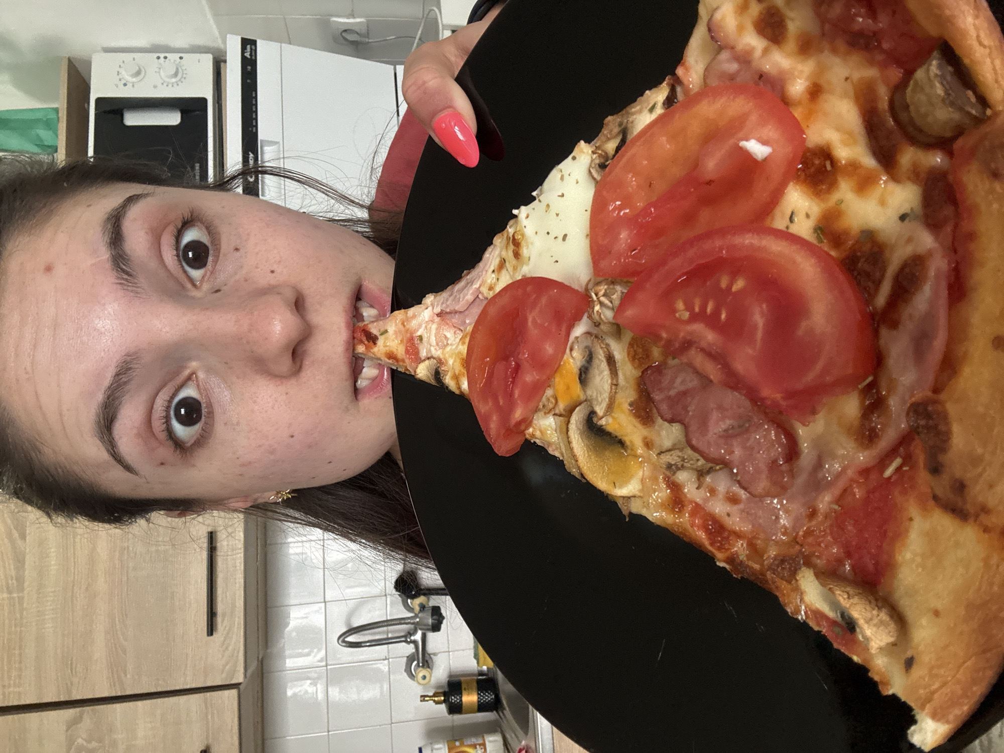 A woman with brown hair and eyes is eating a slice of pizza. The pizza has cheese, tomato, mushrooms and ham. The pizza is on a black plate. The woman has pink nails. The background is a kitchen with a sink and cabinets. The location appears to be indoors.