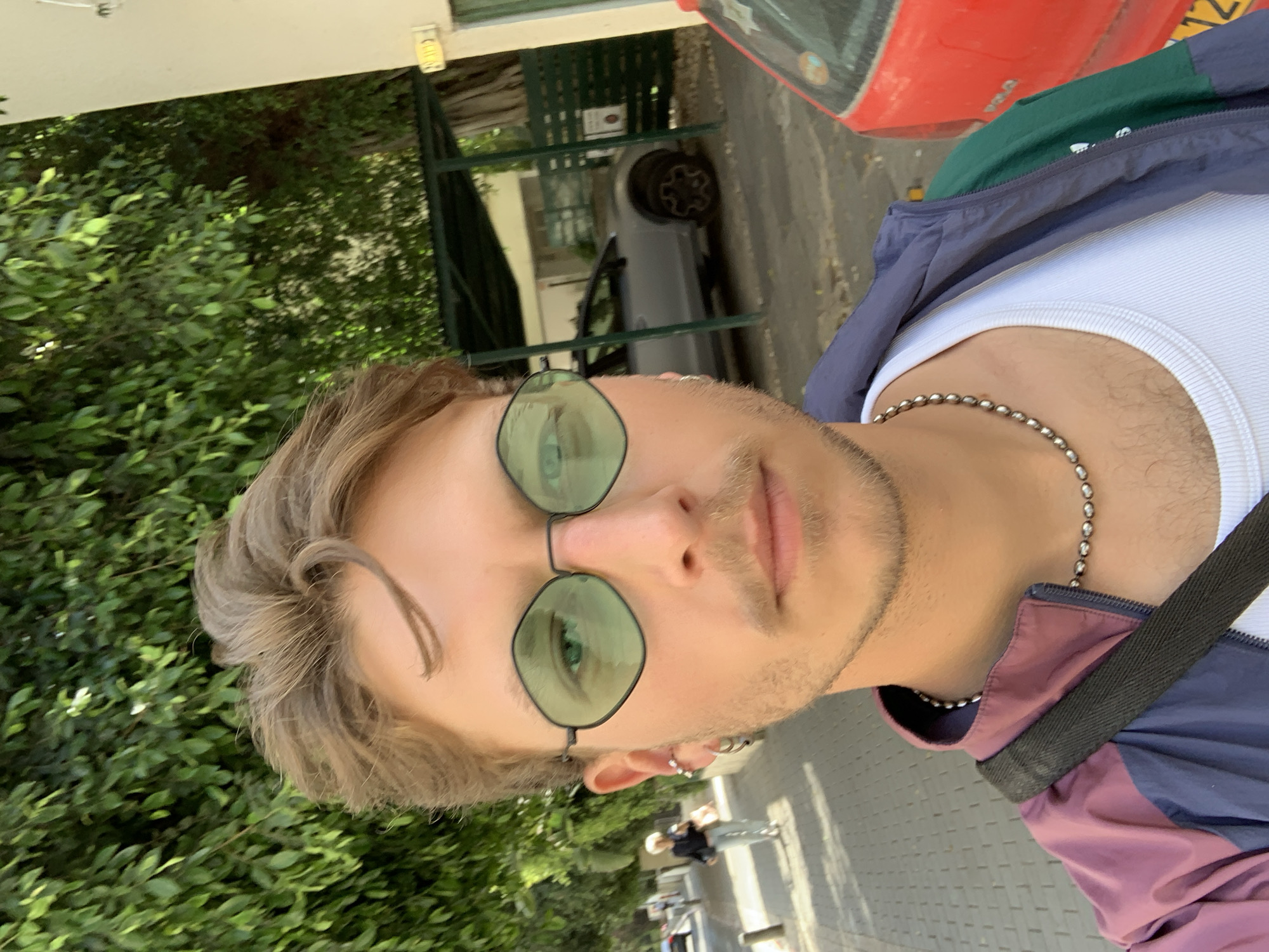 A young man with fair skin, light brown hair, and green hexagonal sunglasses stands outdoors. He wears a white ribbed tank top and a colorful windbreaker. A silver chain necklace adorns his neck. He is facing the camera with a slight smirk.
The background consists of green foliage, a parked silver car, a red vehicle, and some street furniture and pedestrians. This photo was taken on a Tel-Aviv street.