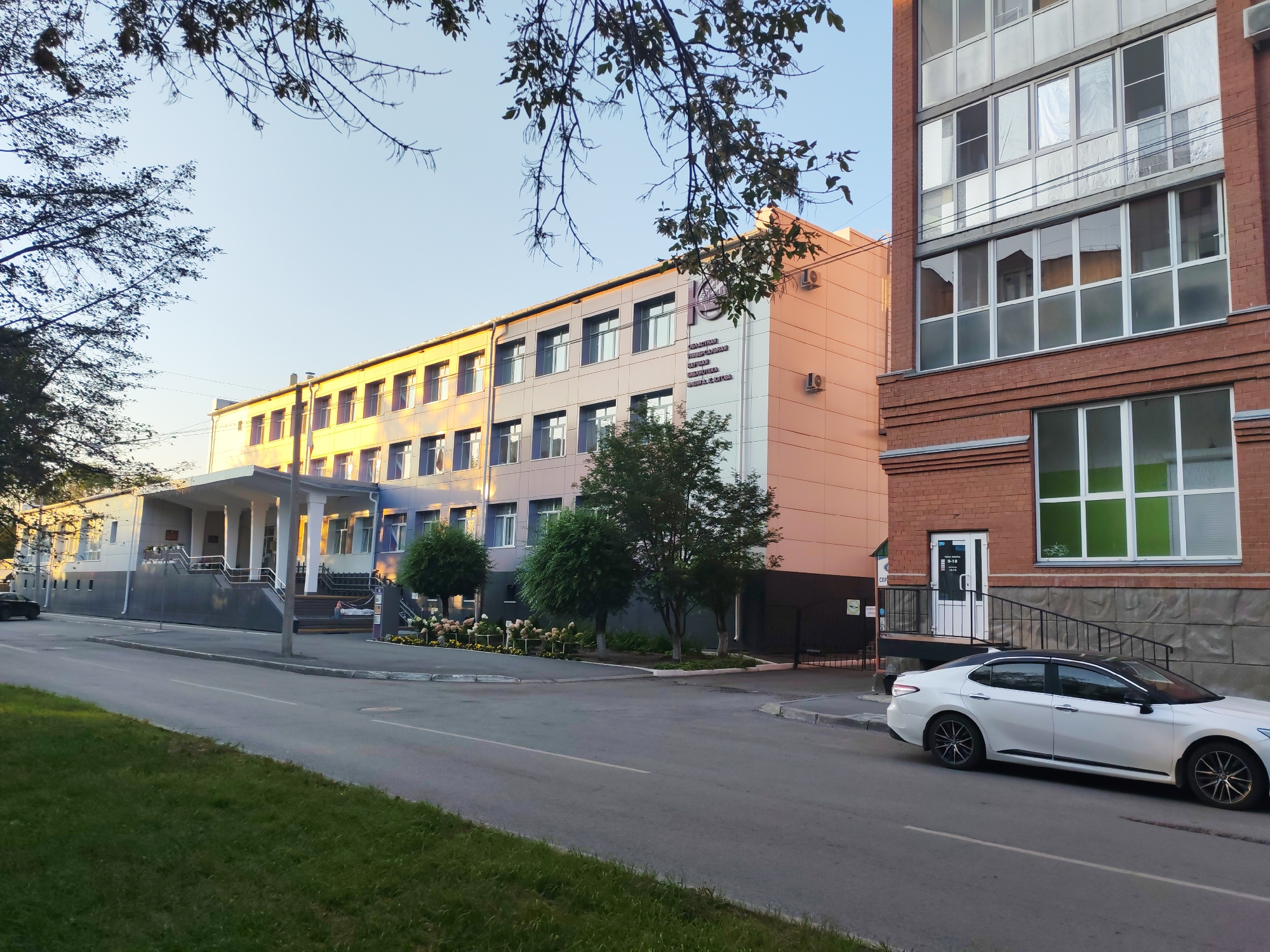 The image shows a modern building with a light-colored facade and a dark-colored roofline, featuring numerous windows. To the right, a brick building with large windows is visible. A white sedan is parked on the street in front of the brick building. The scene is set in an urban environment, with a paved road and a grassy area in the foreground. Trees and landscaping are present around the buildings. The sky is clear and blue, suggesting daytime.