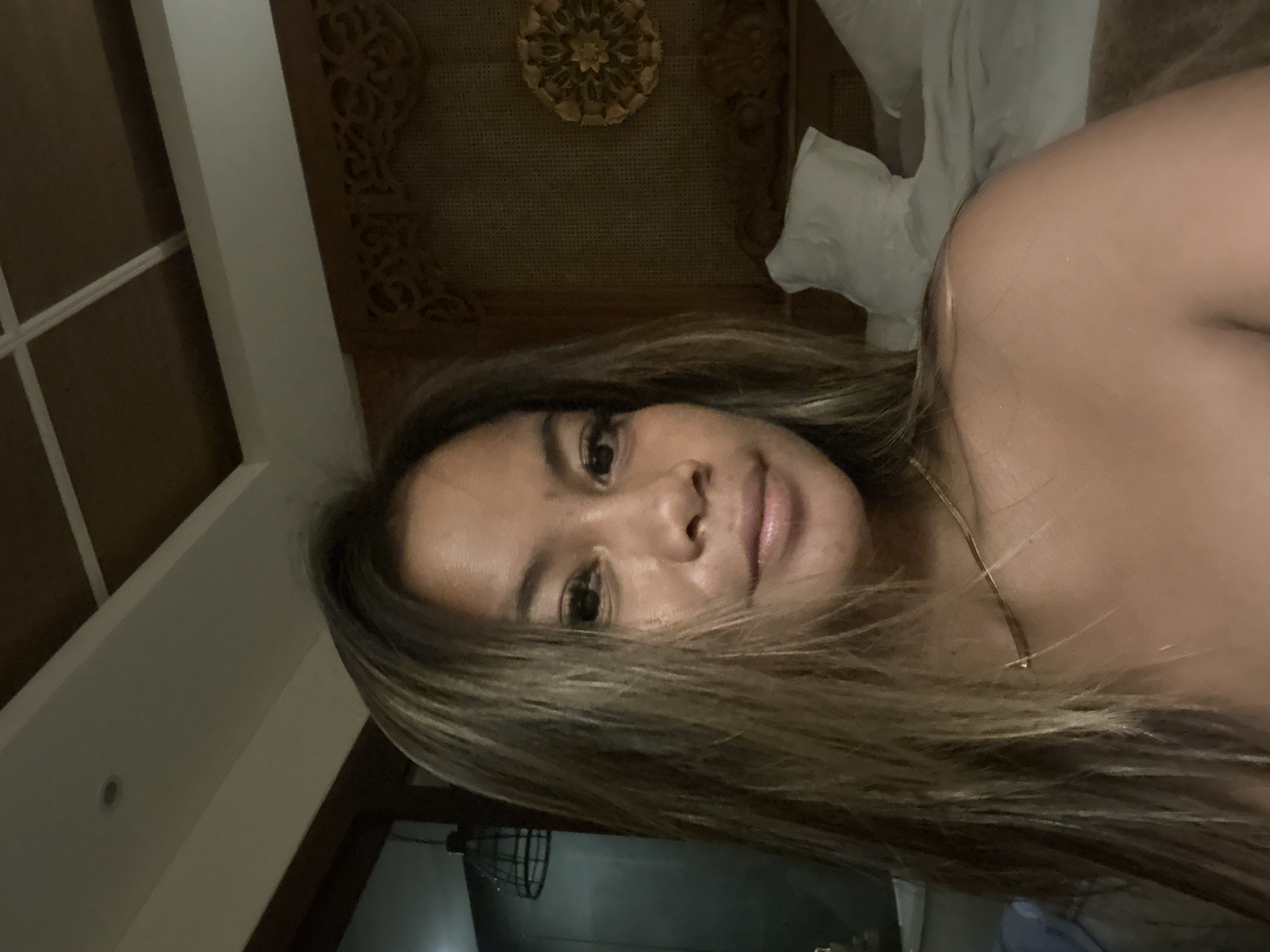 A woman is facing the camera, indoors, lying down. Her hair is light brown and blonde. She is wearing a thin gold necklace. Behind her is a carved wooden headboard with a decorative rosette in the center. Further back, partially visible, is a window with a metal grate. The location is likely a hotel or residence within Ubud District, Indonesia, based on the typical architectural features of the room.