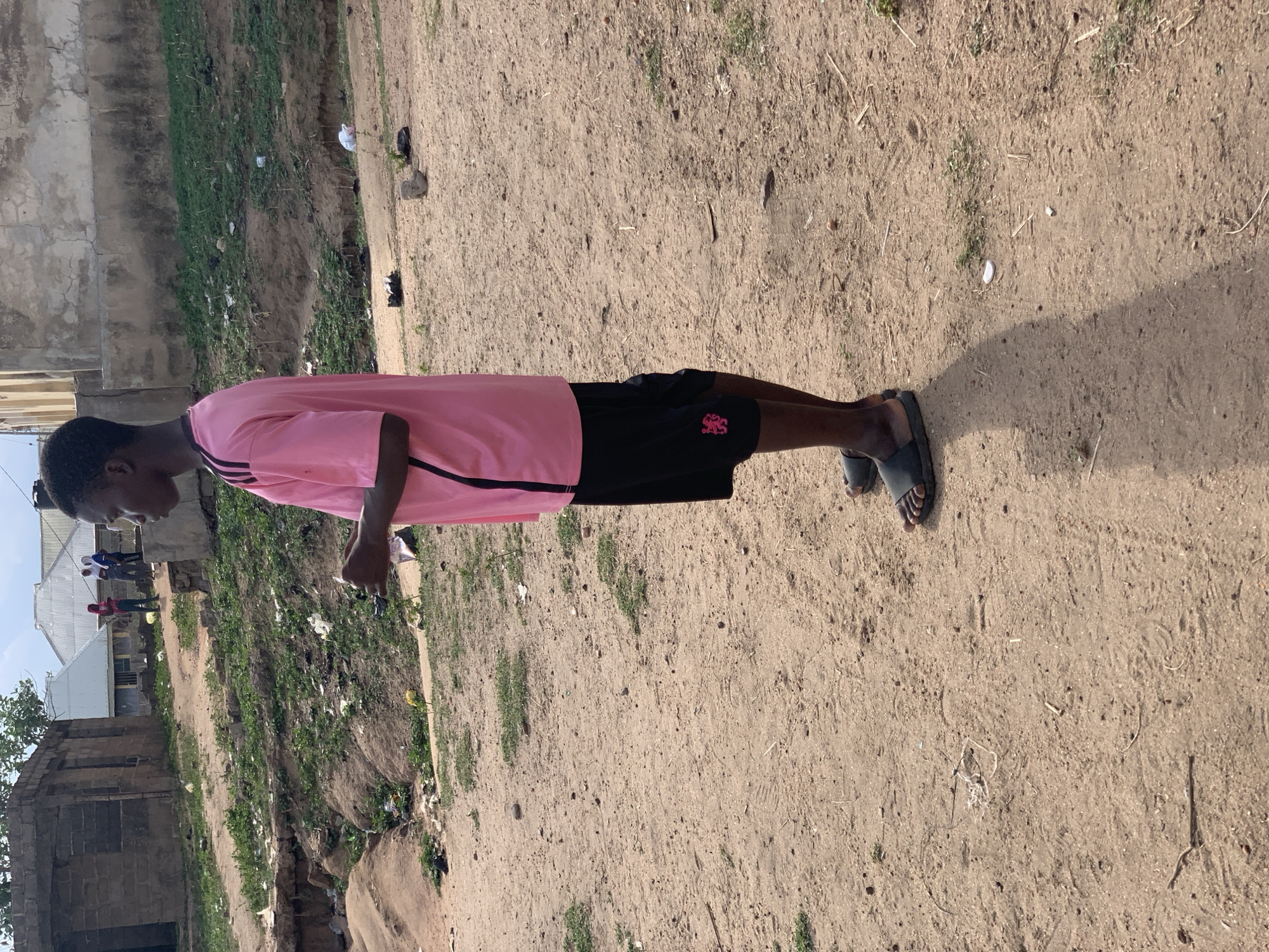 A young person, appearing male, stands on a sandy, earthy ground under clear daylight. The individual is wearing a light pink short-sleeved t-shirt with black stripes on the sleeves, black shorts featuring a pink or red stylized emblem on the left thigh, and dark open-toed sandals. They are positioned in profile, looking towards the left side of the frame with a downward gaze. A distinct shadow extends from their feet to the right, indicating bright sunshine, likely during late morning or early afternoon.
The immediate ground is composed of dry, light brown soil, scattered with small pebbles, dry twigs, and sparse patches of green grass. In the middle ground, the terrain slopes downwards into an overgrown ditch or uneven area, where various pieces of trash, including white and dark plastic debris, are visible. Beyond this ditch, in the background, are several simple, weathered buildings constructed from concrete blocks, some topped with corrugated metal roofs. A few indistinct figures can be seen in the distance near these structures. The overall setting suggests a semi-urban or rural environment, consistent with the provided location context of Lokoja, Nigeria. No discernible text is visible on the clothing or in the surroundings.