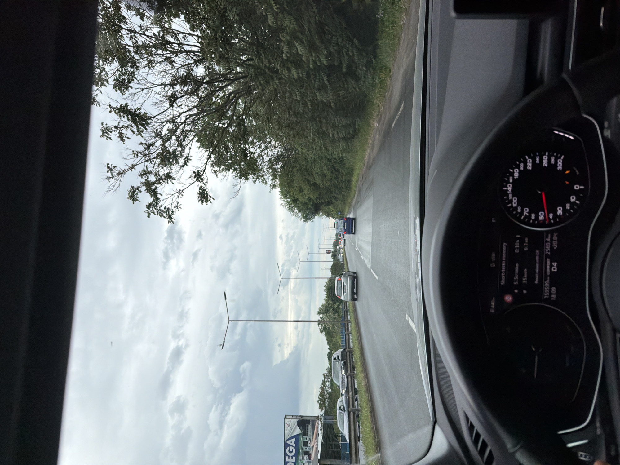 The image is taken from the interior of a vehicle, likely a car, showing the dashboard with the speedometer indicating a speed just below 50 km/h. The digital display on the dashboard shows data including "159599" and "COMFORT 2560.4". The outdoor view through the windshield reveals a road with multiple lanes and other vehicles traveling in the same direction. Street lights line the side of the road, suggesting a highway or major thoroughfare. A billboard with the text "DEGA" is visible to the left of the road. Based on the road signs and the presence of "DEGA", it is likely the image was taken in Burgas, Bulgaria. The environment consists of an overcast sky and green trees lining the road.