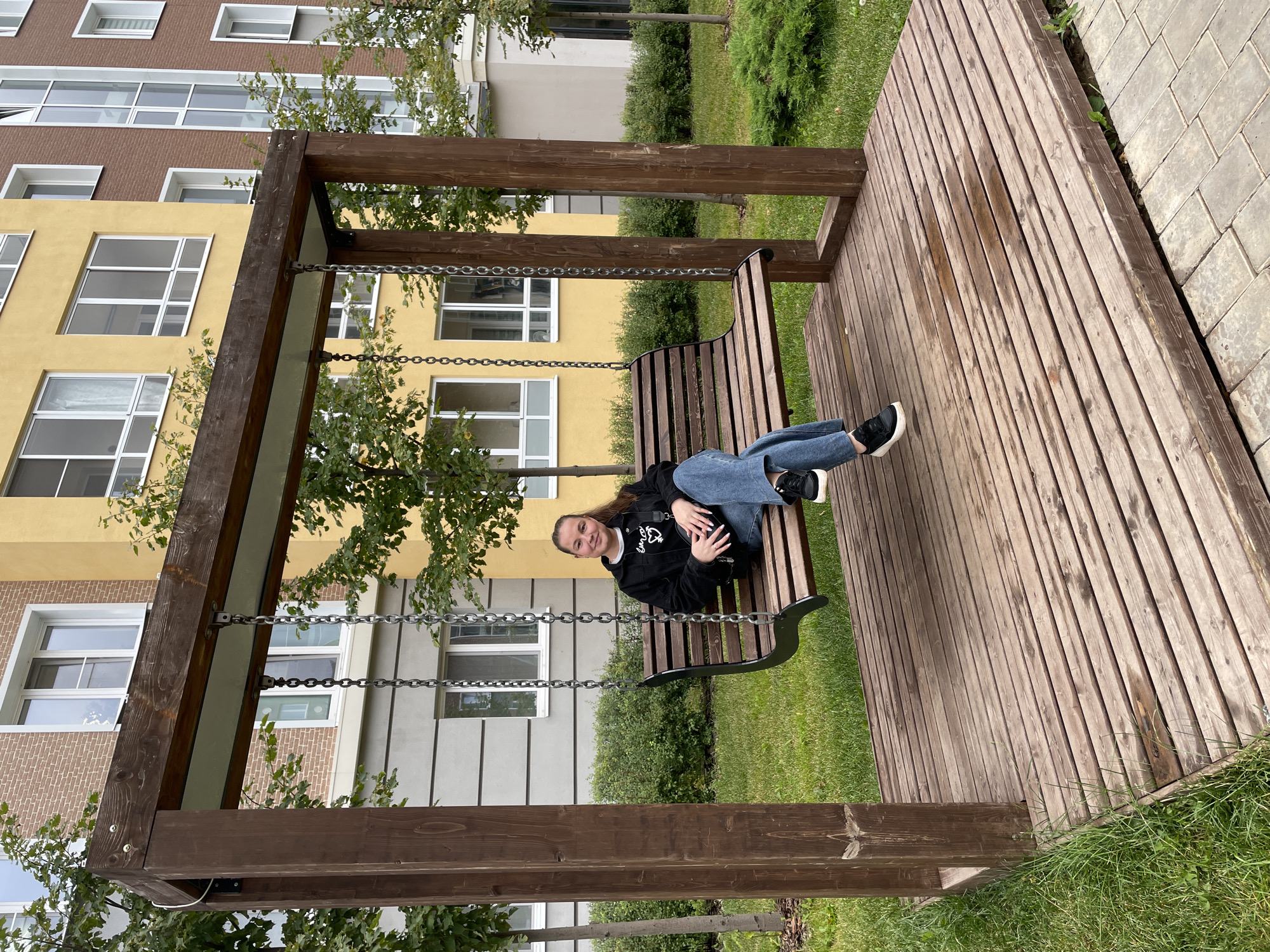A young woman with dark hair pulled back is sitting on a wooden swing in an urban courtyard in Pervomaysky, Russia. She is wearing a black jacket and blue jeans, and she is looking at the camera with a slight smile. The swing is made of dark wood and has a slatted seat. It is suspended from a wooden frame by chains. The ground beneath the swing is a wooden deck. In the background, there are multi-story buildings with yellow and brick facades, as well as green trees and bushes. The overall impression is of a modern residential area with green spaces.