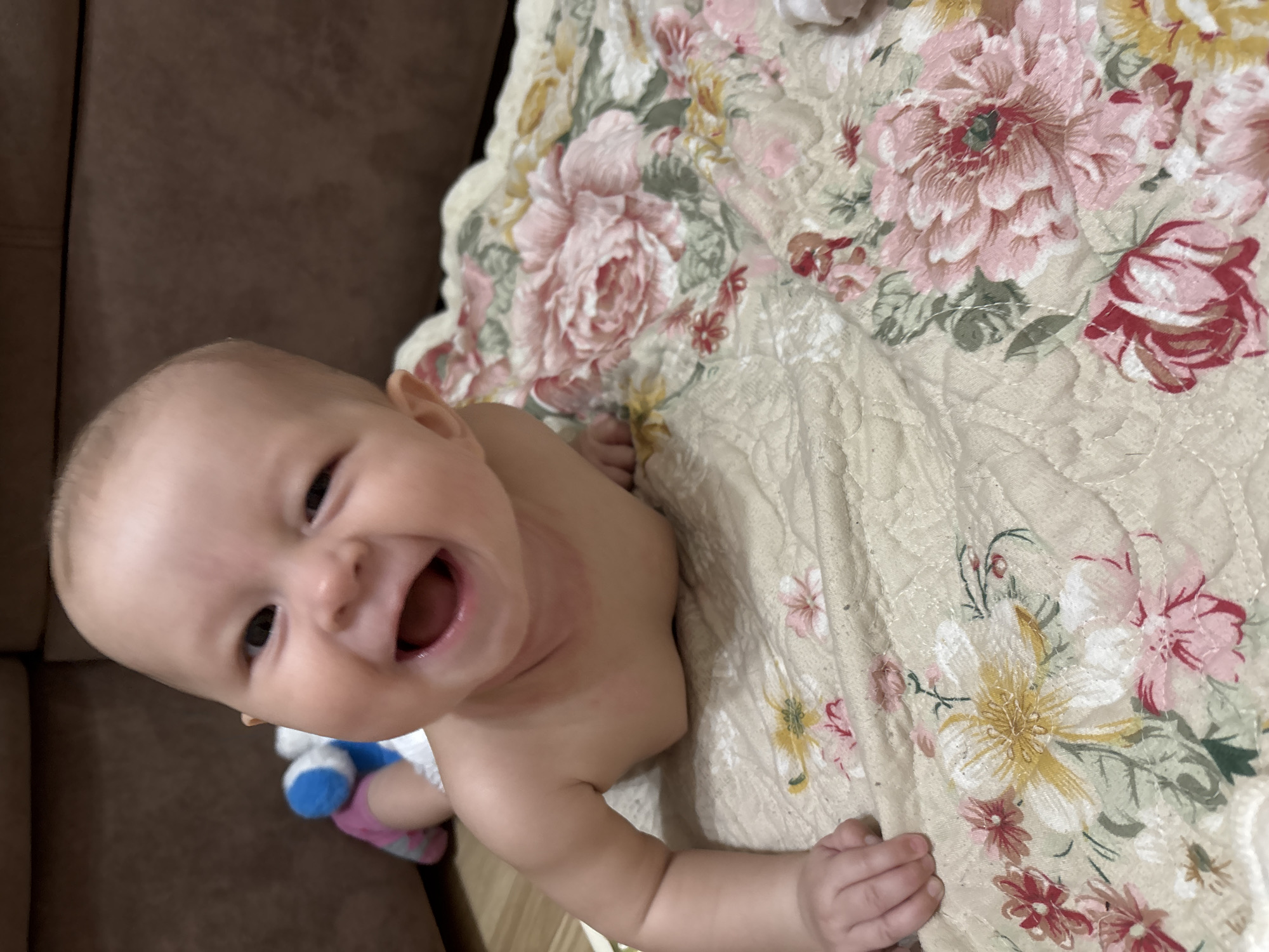 A close-up, angled view shows a baby lying on a floral-patterned quilt. The baby is fair-skinned, has dark eyes, and is smiling broadly, with its mouth open and teeth visible. The baby is wearing a diaper and appears to be on its stomach, with one arm extended and its hand visible on the quilt. A stuffed toy is visible near the baby's legs. The background includes a brown couch. The location is unspecified; the city of Sochi, Russia, cannot be determined from the visual information.