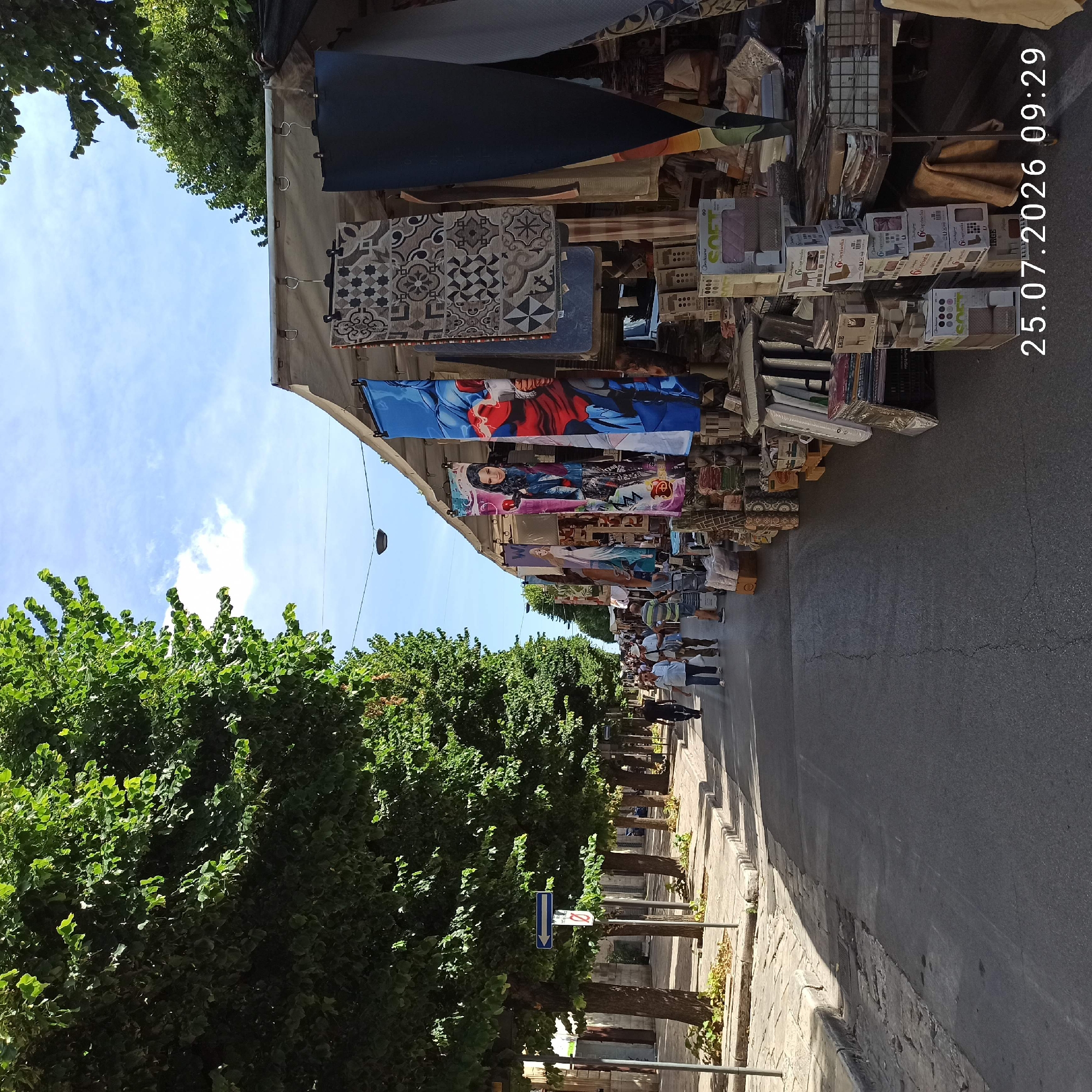 The image depicts a street market in Ruvo di Puglia, Italy, on July 25, 2026, at 09:29. The scene is captured from a street-level perspective, looking down a paved street lined with trees on the left and market stalls on the right.
On the left side of the street, a sidewalk with a stone curb is visible, bordered by mature trees with dense green foliage, casting shadows on the sidewalk and road. A traffic sign with a blue arrow pointing left and a red circular sign with a diagonal red bar are mounted on poles along the sidewalk. People are seen walking on the sidewalk in the distance.
The right side of the street is occupied by market stalls covered by awnings. The stalls display various items, including textiles with patterned designs and large banners featuring animated characters like Superman and female figures. Boxes of products, some stacked and labeled, are visible in front of the stalls. A metal cart with wheels is present, suggesting goods are being moved.
Several individuals are walking along the street and sidewalk. Two men are walking in the foreground on the right sidewalk. In the background, a group of people are seen browsing at the market. The sky is blue with scattered white clouds. The overall atmosphere suggests a bustling outdoor market on a sunny day.