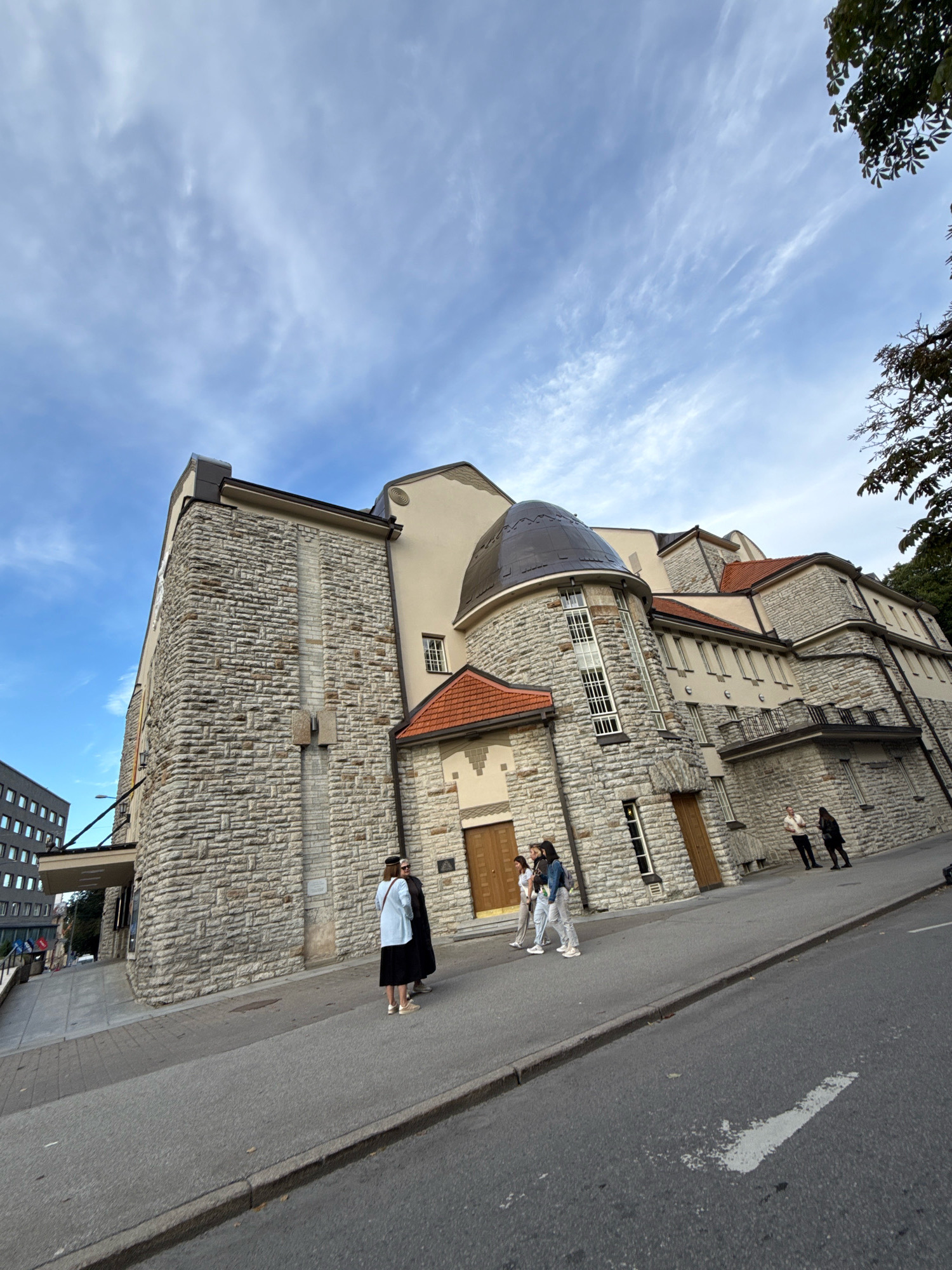This is a low-angle, wide shot of the Vabamu Museum of Occupations and Freedom in Tallinn, Estonia, on a cloudy day. The stone building features a distinctive domed roof section and a tiled roof. Several people are visible on the sidewalk in front of the museum. On the left, two individuals stand near the entrance, one wearing a light blue jacket and a black skirt, the other in a black coat. Further to the right, a group of three young people in casual attire are walking. In the background, two more figures are standing near another doorway. The street in the foreground has a white arrow painted on it. The sky is partly cloudy with wispy cirrus clouds. A portion of a modern, gray building with many windows is visible on the far left edge of the frame.