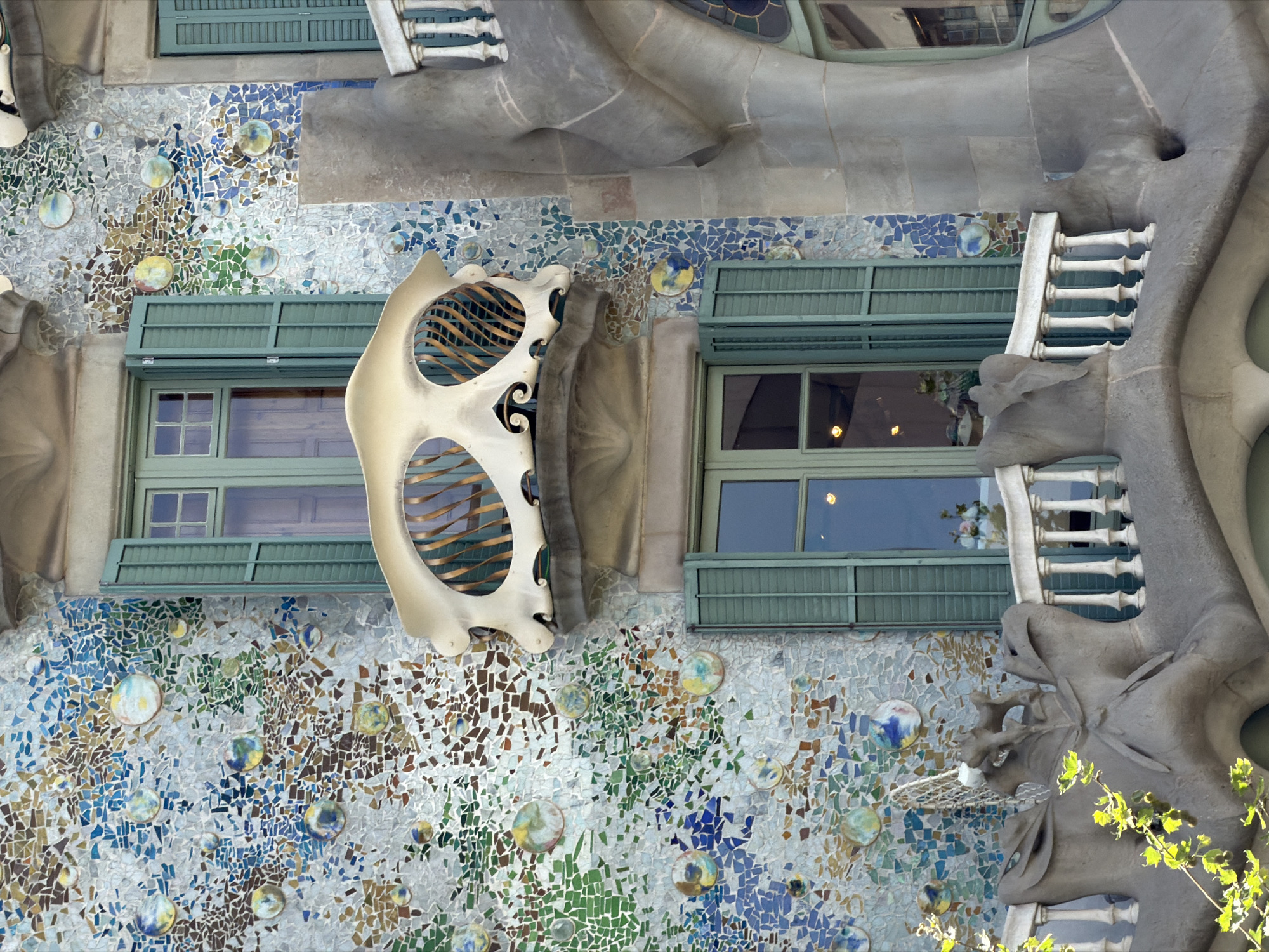The image shows a detail of the facade of Casa Batlló, a building designed by Antoni Gaudí, located on Passeig de Gràcia in Barcelona, Spain. The facade is adorned with a mosaic of colorful tiles and ceramic pieces, featuring a whimsical, organic design. Two balconies are visible. The upper balcony has a unique, bone-like white structure with an opening filled with decorative metalwork. Below it, another balcony features a white balustrade supported by sculpted, gray stone elements that resemble organic forms. Green shutters are open on the windows flanking the upper balcony, and the windows below are also visible. The overall impression is one of vibrant color and imaginative architectural detail.