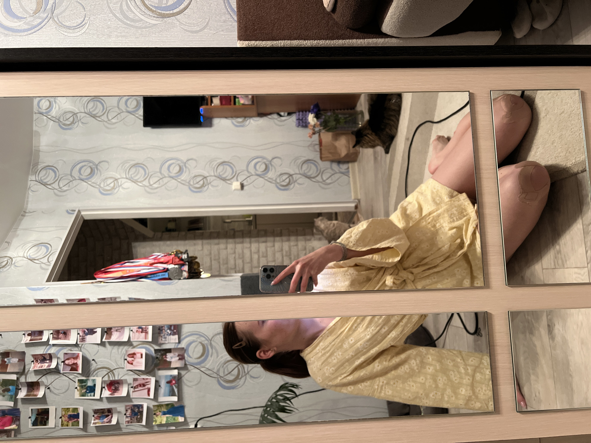The image shows a reflection in a multi-panel mirror, likely in a domestic setting. The mirror reflects a room with patterned wallpaper, a television, shelves, and various objects, including a red and white toy boat. A person is holding a phone, seemingly taking a picture of themselves in the mirror. The reflection captures their upper body, wearing a light yellow floral dress. Also reflected is a cat, and a collection of photographs attached to the wall. The top portion of the mirror reflects what appears to be a person's legs and feet. There is no evidence to confirm the location is Chaikovsky, Russia.