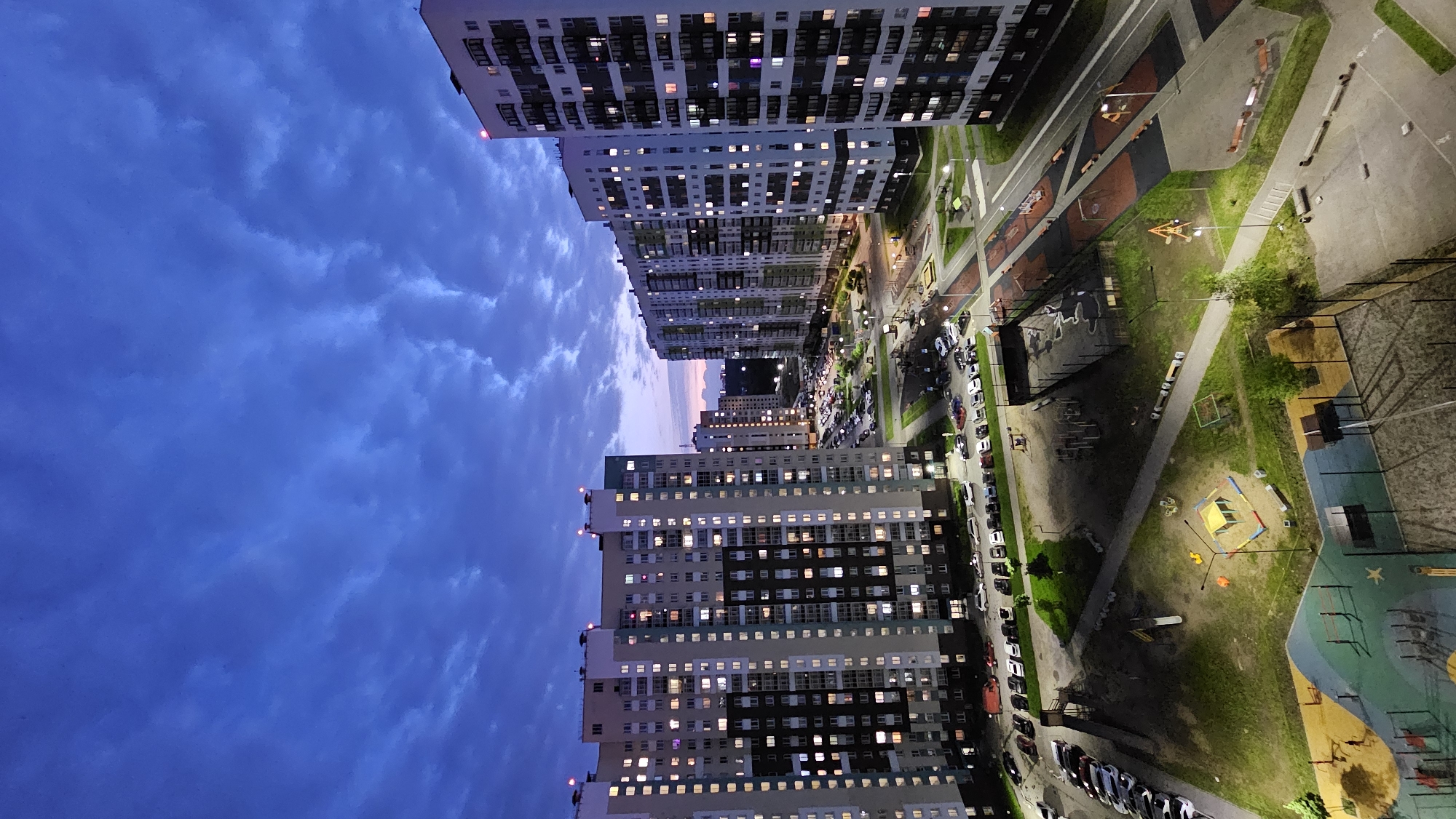 This is an aerial, evening photograph of a residential complex in Yekaterinburg, Russia. The sky is a deep blue with scattered clouds, indicative of twilight. Several tall, modern apartment buildings dominate the scene, their windows illuminated with warm light, suggesting that residents are home. Parking lots filled with cars are visible between the buildings, and well-maintained playgrounds with various equipment, including a sandbox, swings, and slides, are scattered throughout the complex. The ground is covered in grass and paved pathways. The overall impression is one of an organized and active urban living environment.