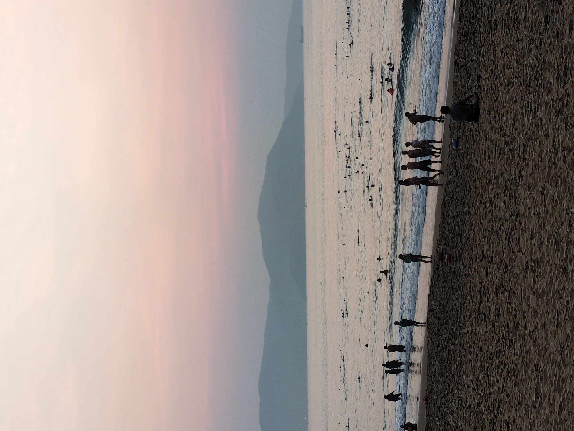 The image depicts a coastal scene in South Quy Nhon Ward, Vietnam at either sunrise or sunset. The sky shows a gradient from pale pink near the horizon to a lighter, more faded tone above. A mountain range is visible in the distance, appearing hazy and pale blue. The ocean occupies the central portion of the image, with many objects and people swimming in the water near the waves. The beach is a dark sand with several groups of people standing at the shoreline. One person is sitting on the sand.