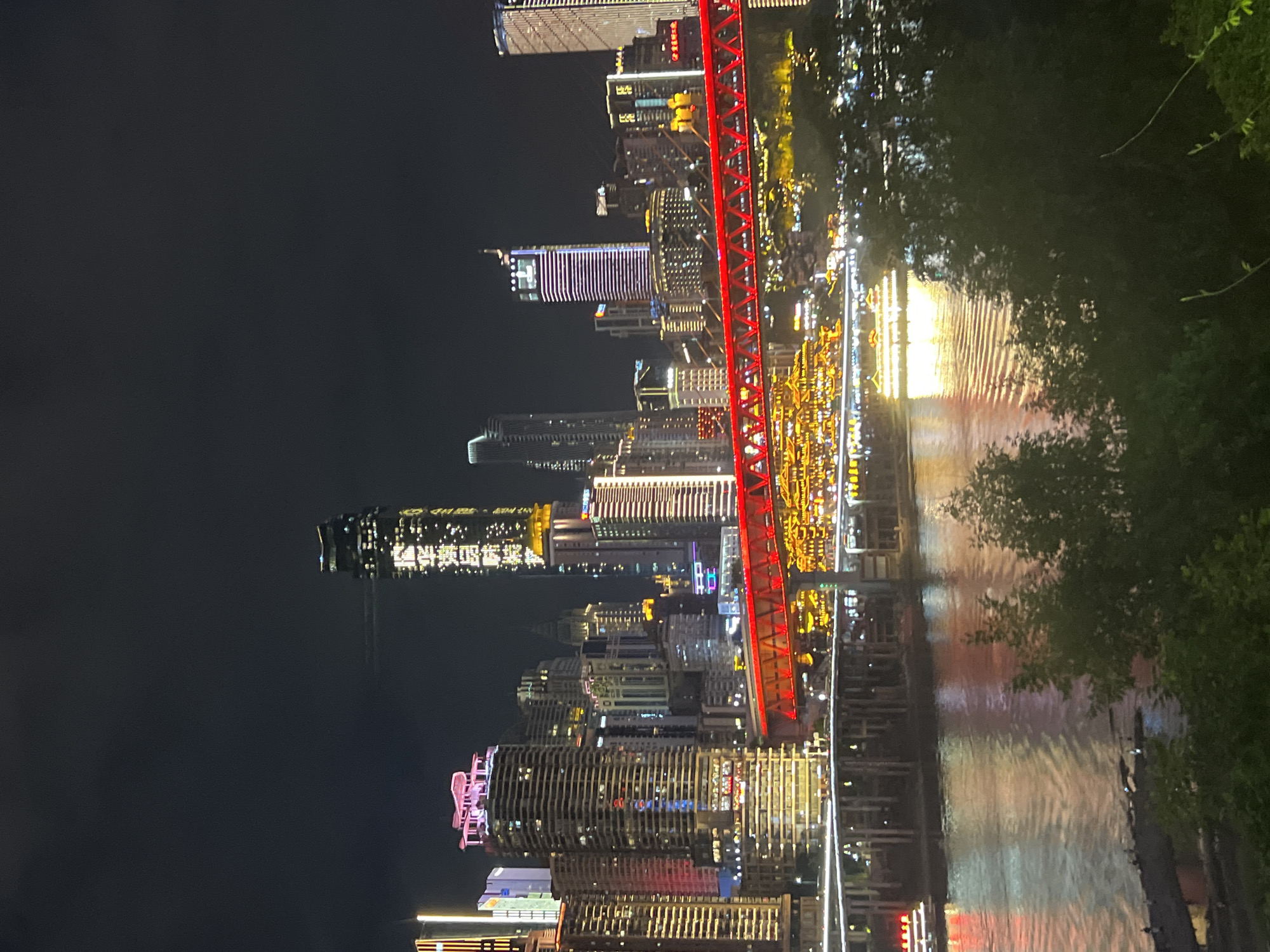 The image depicts a nighttime cityscape of the Jiangbeicheng Street area in China, characterized by a dense concentration of illuminated skyscrapers and a prominent red bridge spanning a body of water. The buildings, varying in height and architectural style, are adorned with numerous lights, creating a vibrant and complex urban panorama. A notable structure on the right is a tall skyscraper with vertical purple lighting and another on the left with horizontal yellow and pink lighting. The red bridge features a distinctive lattice design and is illuminated from below and along its structure, casting a warm glow on the water. The water reflects the city lights, creating shimmering patterns on its surface. A large, illuminated boat is visible on the water, adding to the lively atmosphere. The foreground is dominated by dark, silhouetted foliage, suggesting a vantage point from a hill or elevated area overlooking the city and river. The sky is dark, emphasizing the artificial illumination of the urban environment.