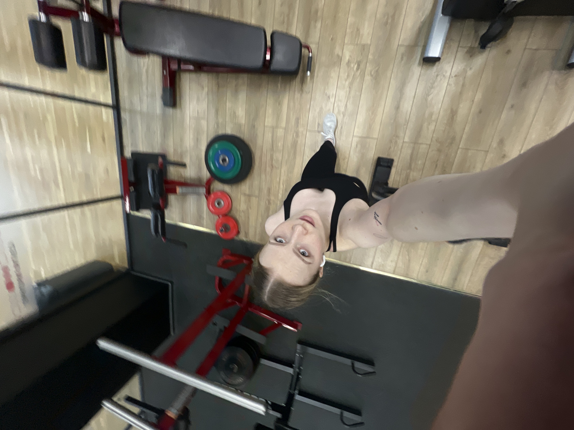 A woman is taking a selfie from a high angle in a gym. She is wearing a black sports bra and leggings, with a white earbud in her ear. The gym appears to have standard weightlifting equipment, including a bench press, weight plates, and squat rack. The floor is a light-colored wood laminate in one area and a black rubber material in another. The location is in Khimki, Russia.