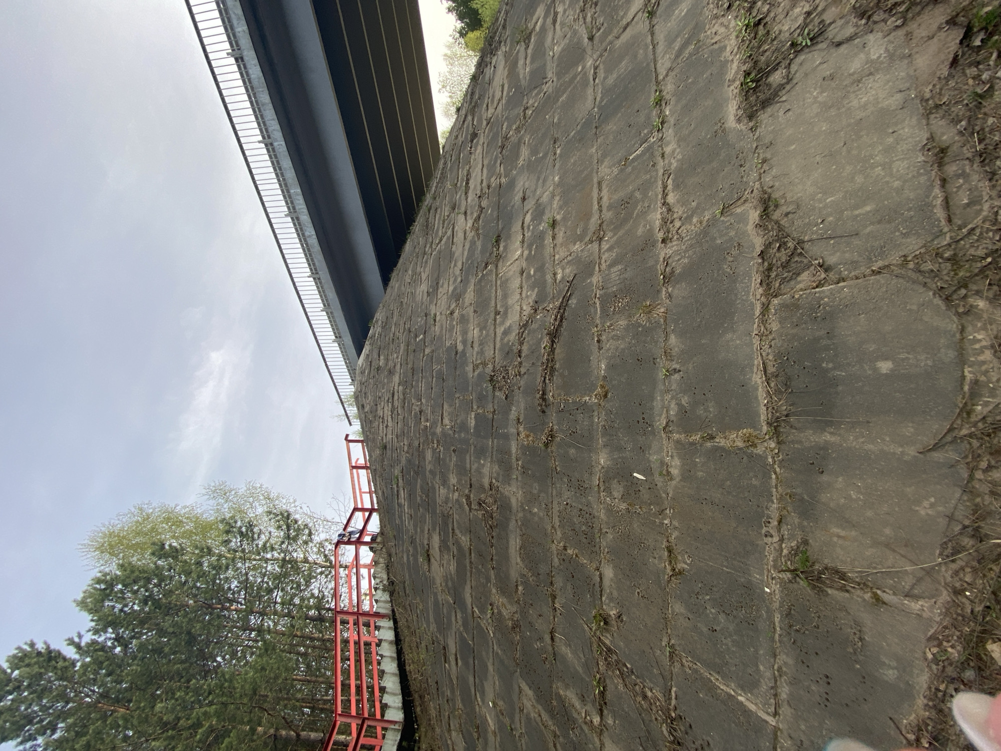 The image shows a view of a concrete wall, likely a retaining wall or part of a bridge abutment, located in Chaikovsky, Russia. The wall is constructed of large concrete blocks with visible joints and is covered with some vegetation and debris. Above the wall is a section of a bridge or overpass with a guardrail. To the left of the wall are trees and a red metal structure, possibly a staircase or access platform. The sky is overcast. There are no people visible in the image.
