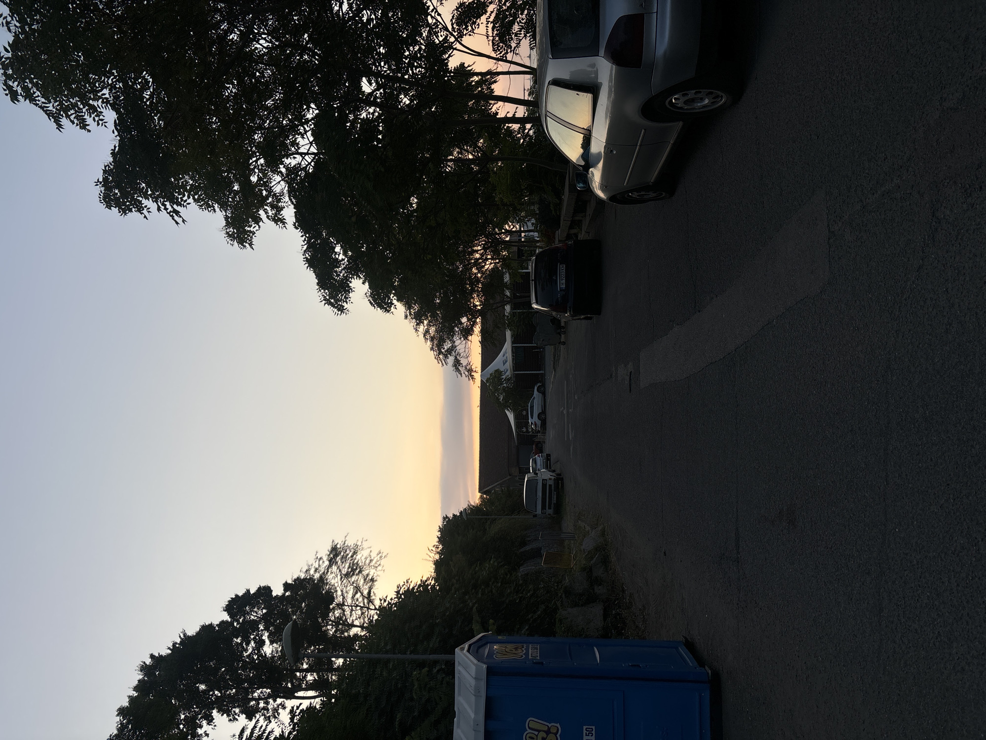 The image captures an outdoor scene at dusk, likely in Tsarevo, Bulgaria, given the context. A paved road leads into the distance, flanked by trees and vegetation. On the left, a bright blue portable toilet is visible. Several vehicles are parked along the road, including a silver hatchback, a dark-colored sedan, and a white van. In the background, there are buildings with pitched roofs and white tent-like structures, suggesting a public area or event. The sky displays a gradient of pale blue and soft orange hues, characteristic of twilight. The overall impression is a quiet, late-day ambiance.