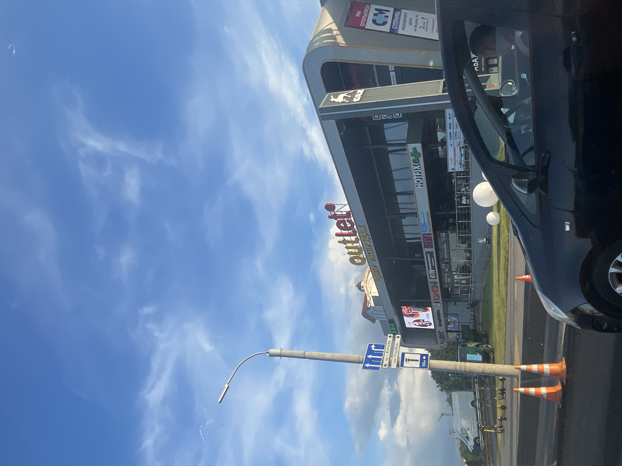 The image shows a street view with a large building in the background labeled "outlet". A street light with directional signs is in the foreground, and traffic cones are on the road. A black car is partially visible in the lower right of the frame. The sky is blue with some clouds. The building appears to be a shopping center with various signs, including one for "HOTFIX Coffee" and another with text that is difficult to read. There are also large white spheres placed on the grass in front of the building. A white van is parked on the left side of the image. The overall scene suggests a commercial area on a clear day.