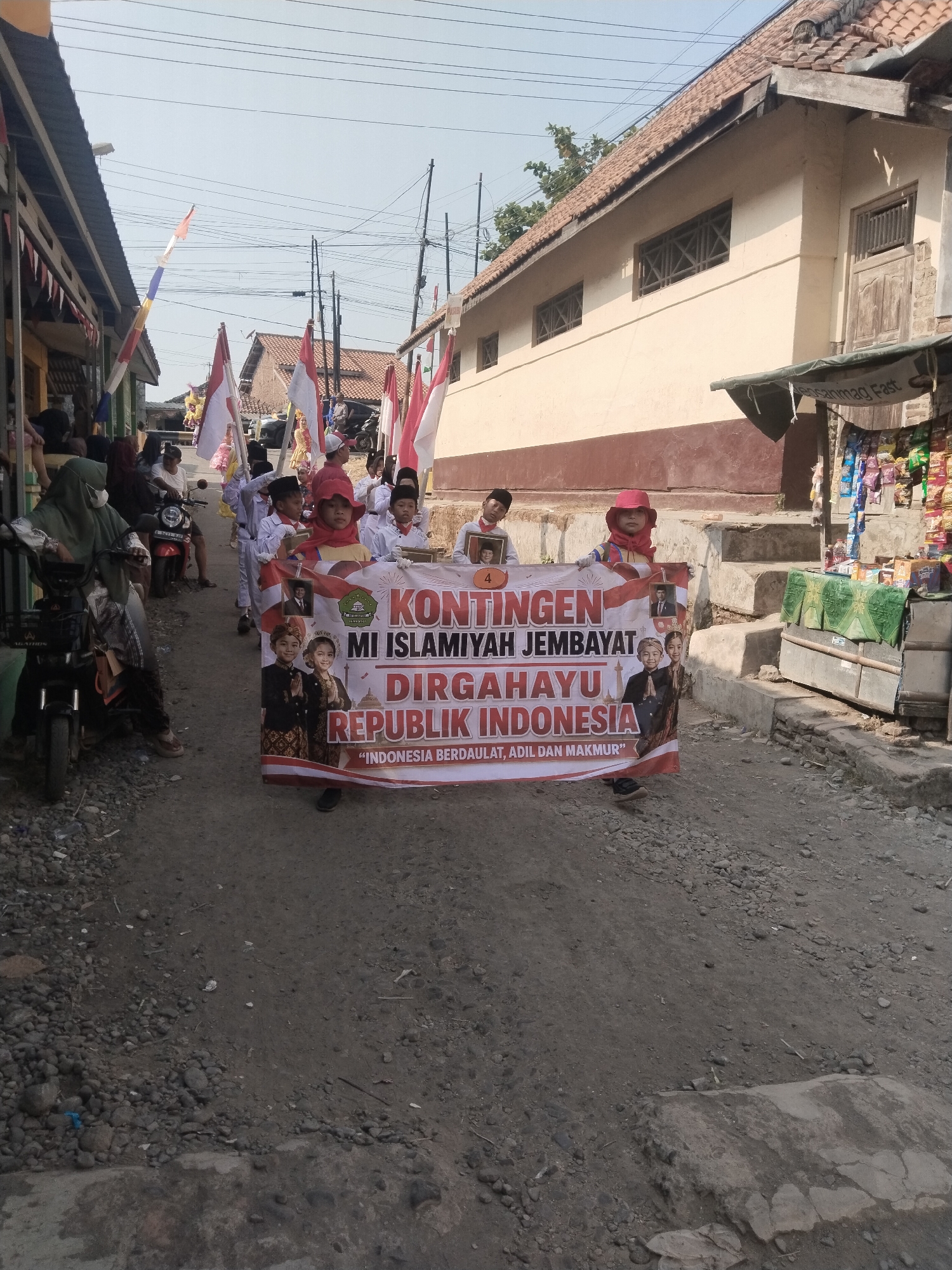 In Jembayat, Indonesia, a group of children in uniform, some wearing red hats and others white hats, are participating in a parade. They are holding a banner that reads "KONTINGEN MI ISLAMIYAH JEMBAYAT DIRGAHAYU REPUBLIK INDONESIA" and "INDONESIA BERDAULAT, ADIL DAN MAKMUR". The parade is taking place on a dirt road lined with buildings and trees. Several red and white Indonesian flags are visible, some attached to poles and others carried by participants. In the background, there are motorcycles and spectators. The scene suggests a celebration or commemoration of Indonesian Independence Day.