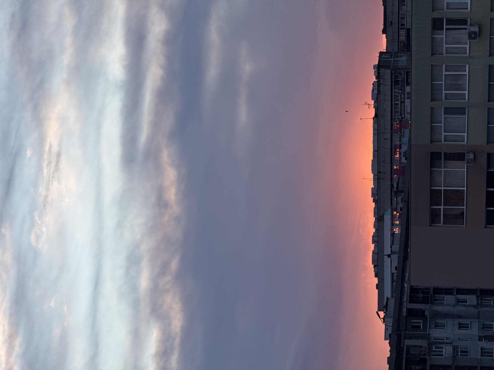 The image depicts a cityscape at sunset. On the right side, tall buildings with multiple windows are visible, some with air conditioning units. The building closest to the viewer is partially obscured by shadows and shows signs of aging. The skyline is sharply defined with the silhouettes of various structures including smaller buildings and antennae. The sunset casts an orange and pink glow behind the buildings, reflecting in some windows. The sky takes up the majority of the frame, displaying a gradient from pale blue to a deeper purple-blue with elongated cloud formations.
Without additional information, it is difficult to confirm the exact location in Burgas, Bulgaria.