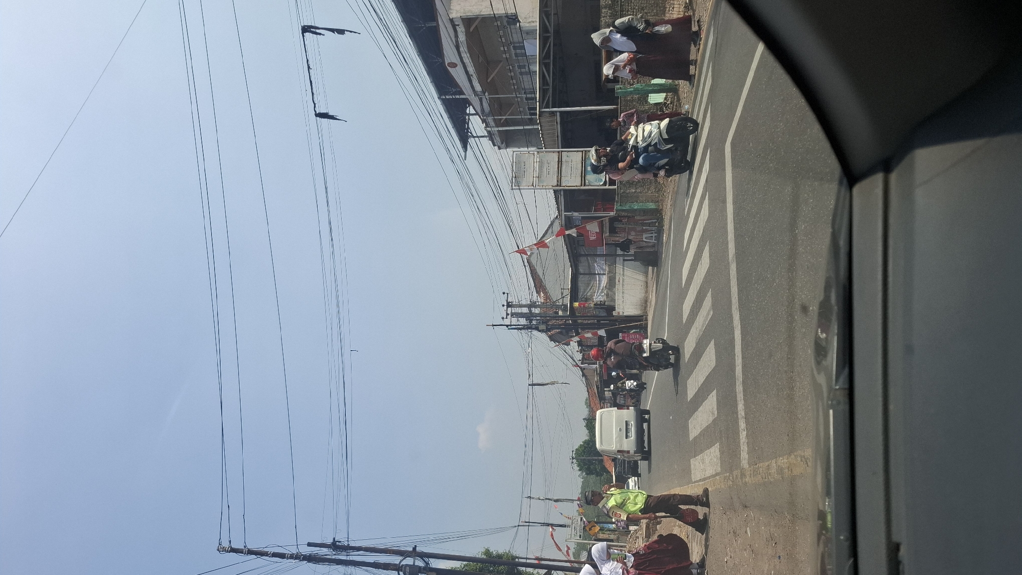 The image captures a street scene in Sirnagalih, Indonesia, likely during daytime, with a clear blue sky. A white van is visible on the road, positioned to the left of a pedestrian crossing. Several individuals are present near the crossing. On the left, two people, one wearing a green vest, stand on the side of the road. To the right of the pedestrian crossing, a person rides a white scooter, accompanied by a passenger. Further to the right, three people, two appearing to be wearing hijabs, are standing on the sidewalk. The road is paved, and there are buildings lining the street, some adorned with Indonesian flags. Electrical wires are strung across the scene, supported by utility poles. The foreground shows the interior of a vehicle, likely the perspective from which the photo was taken.