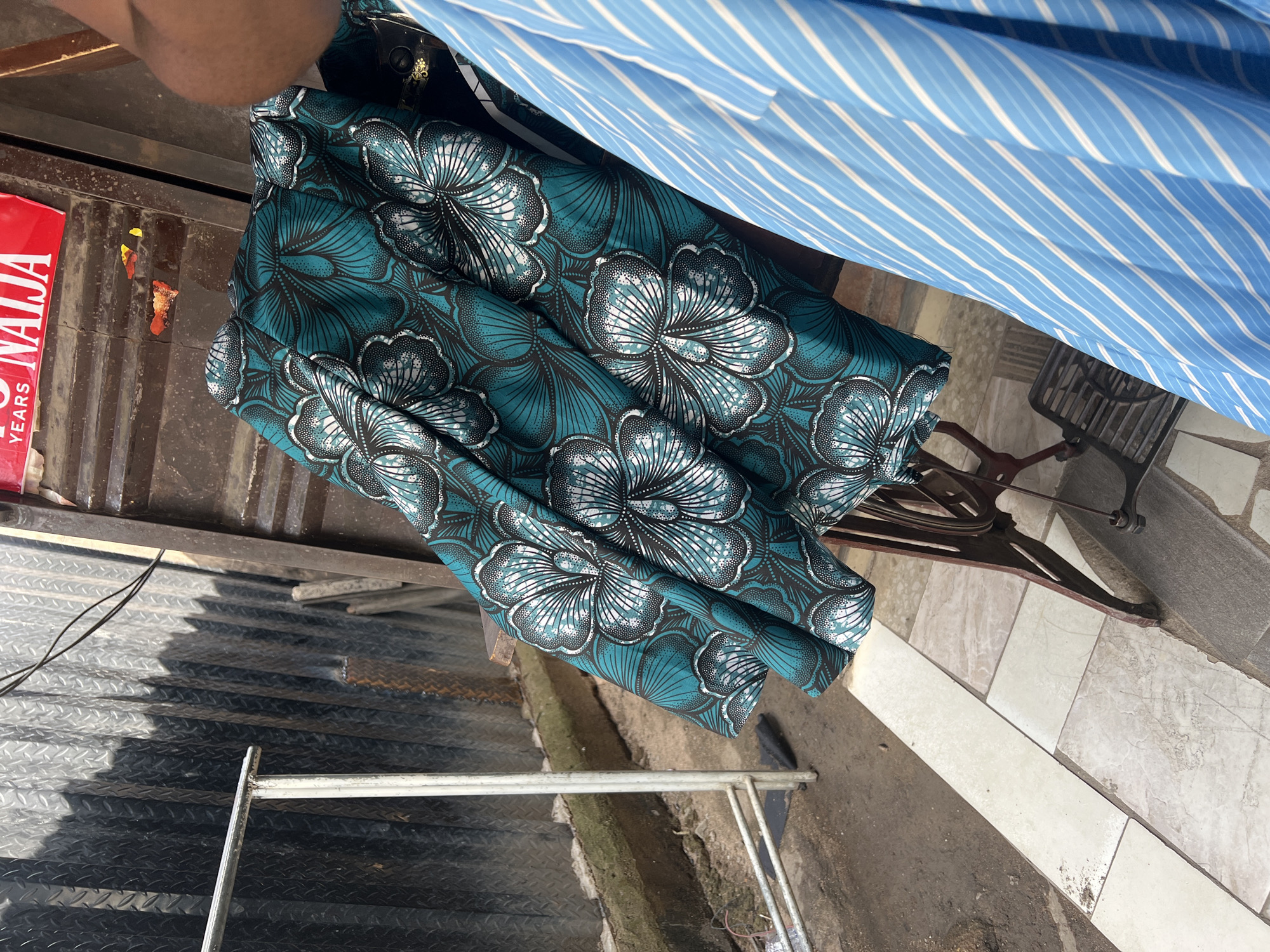 The image captures a close-up view of fabrics laid out on an antique sewing machine, likely in a workshop or market stall in Jalingo, Nigeria. A woman, partially visible from the shoulder up, is in the foreground, wearing a blue and white striped garment. The main subject is a vibrant teal fabric with a bold floral pattern in white and black, draped over the sewing machine. The sewing machine itself is dark and appears to be made of metal with decorative elements.
The environment suggests an outdoor or semi-outdoor setting. Behind the sewing machine, a corrugated metal wall, possibly part of a shop or building, is visible. The ground is paved with large, square tiles of varying shades of beige and gray, with some areas of dirt and debris. A metal clothing rack stands to the left, and some wires are also visible, suggesting a functional workspace. A red sign with white text, partially obscured, is affixed to the metal structure in the background. The lighting indicates it is daytime, with shadows cast by the objects, highlighting the textures and folds of the fabrics.