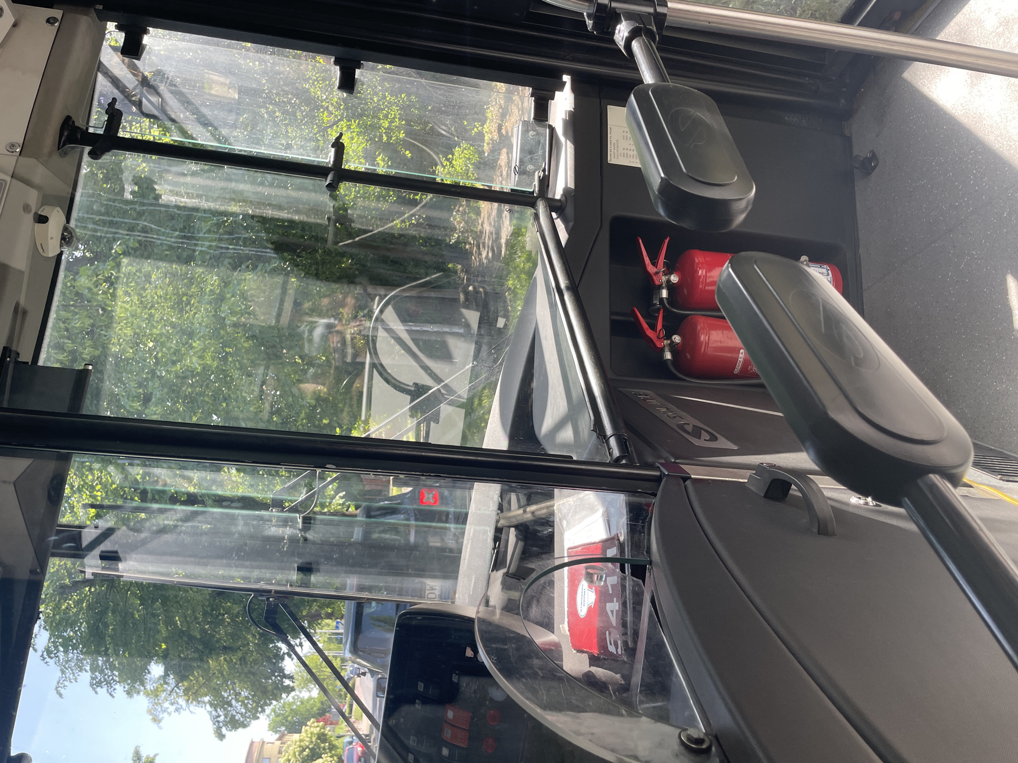 The image shows the interior of a bus, specifically the driver's area, with a view through the windshield to the outside. Two red fire extinguishers are visible to the right of the windshield. The exterior view includes trees and buildings, with other vehicles on the road. The location is Wrocław, Poland.