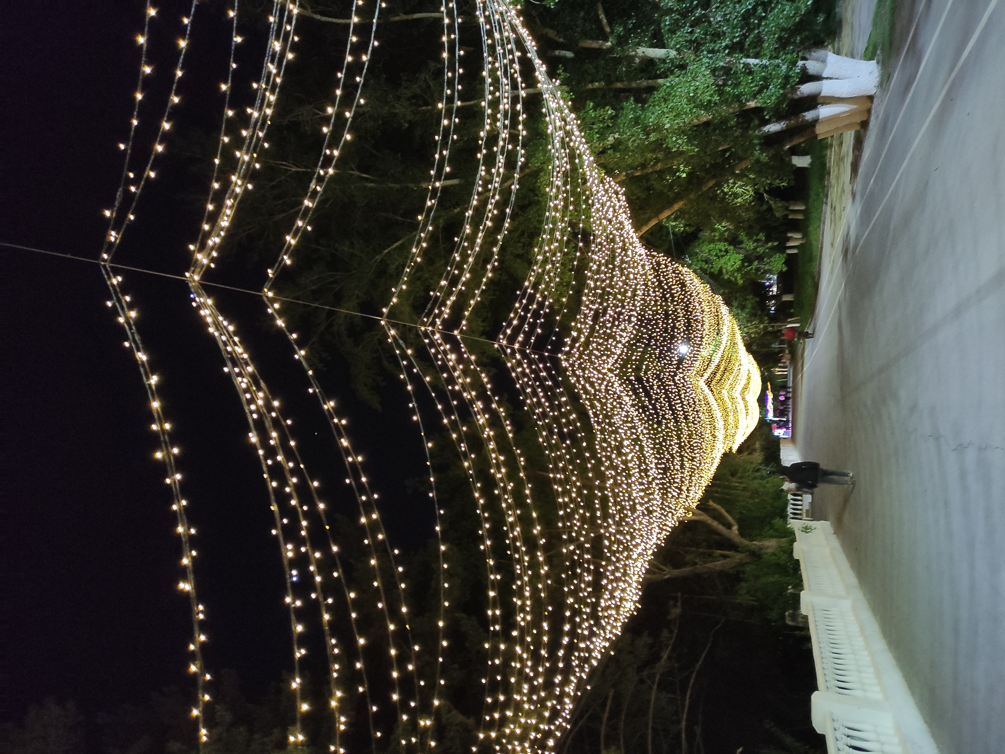 The image shows a nighttime view of a tunnel of lights strung over a pathway in Pavlodar, Kazakhstan. The path, made of pavement, is bordered by a white balustrade on the right and trees on both sides. A person is walking along the path under the lights, and other people are visible in the distance. The tunnel of lights creates a bright, concentrated illuminated space, contrasted by the surrounding darkness.