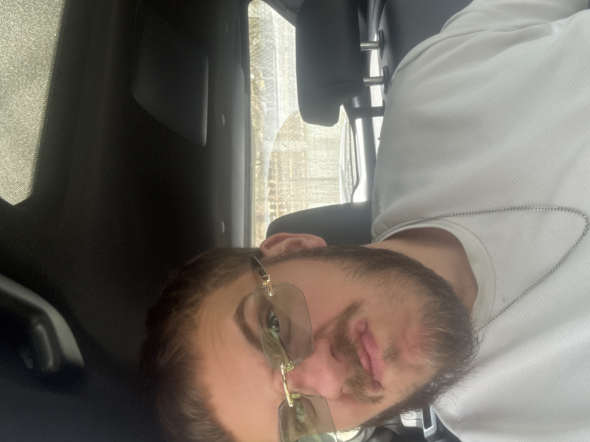 A selfie, oriented sideways, shows a man with brown hair, a beard, and gold-framed sunglasses. He is wearing a white shirt and a silver chain around his neck. The photo appears to have been taken inside a vehicle, based on the headrest behind him and the car's interior elements visible. The location is presumably in Teteven, Bulgaria. The man is looking toward the camera. The scene shows the interior of the vehicle, with a glimpse of an outside environment through the car window.