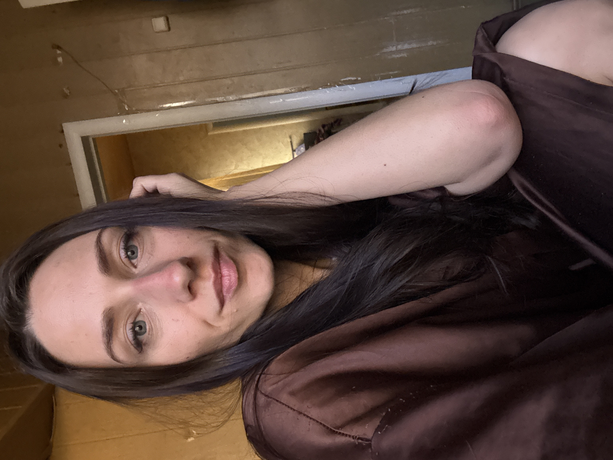 The image features a young woman reclining indoors, captured in Saint Petersburg, Russia. She is seen from the chest up, with her head slightly tilted and turned towards the viewer, displaying a neutral to faintly smiling expression. Her long, dark hair frames her face and spreads around her shoulders. She has green eyes, well-defined eyebrows, and light pink lips. Her left arm is bent, with her elbow visible and resting on a surface, showing a slight reddish mark. She is wearing a dark brown or maroon garment, possibly a robe or top, made of a shiny, satin-like material that drapes around her.
The background shows an interior space with a rustic or aged aesthetic. A wall constructed from light brown wooden planks or panels is visible behind her, characterized by a textured and somewhat worn appearance. To her left (viewer's right), a white-framed doorway leads into another room, which is dimly lit and contains indistinct shapes of furniture or objects. The lighting in the scene is soft and diffused, indicating an indoor setting without direct harsh light. No visible text is present in the image.