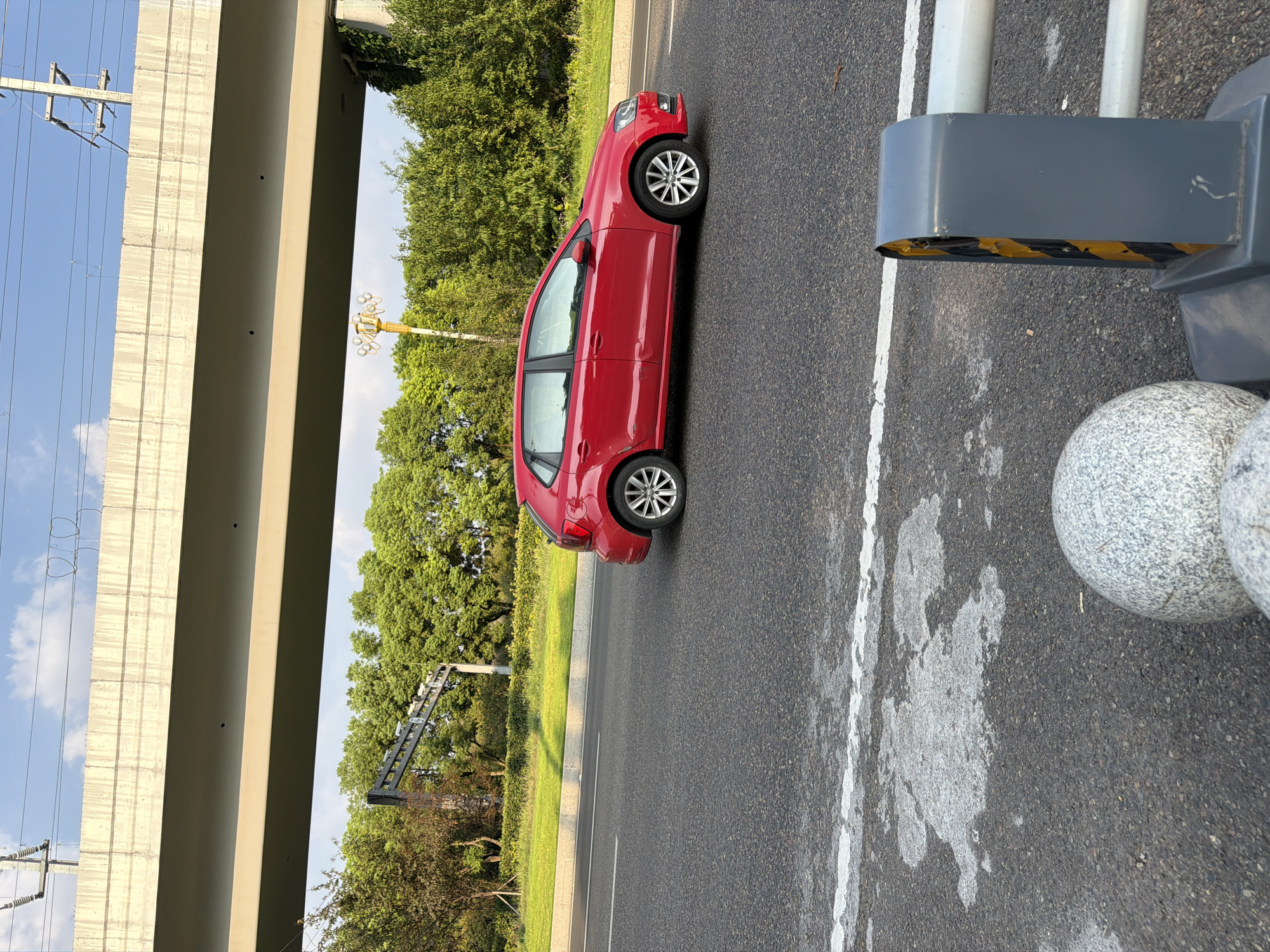 A red Volkswagen Polo hatchback is captured in motion on an asphalt road, illuminated by natural daylight under a clear blue sky. The car is positioned in the right lane, with its right side facing the camera. Behind the car, a lush green landscape with dense trees and foliage is visible, suggesting a park or natural area bordering the road. Overhead, a concrete overpass casts a shadow across the scene, and power lines are strung between utility poles. In the foreground, the road is marked with white lines, and a grey traffic barrier with yellow and black hazard stripes stands to the right, next to two spherical granite bollards. The setting appears to be a road in Jiangjin, China.