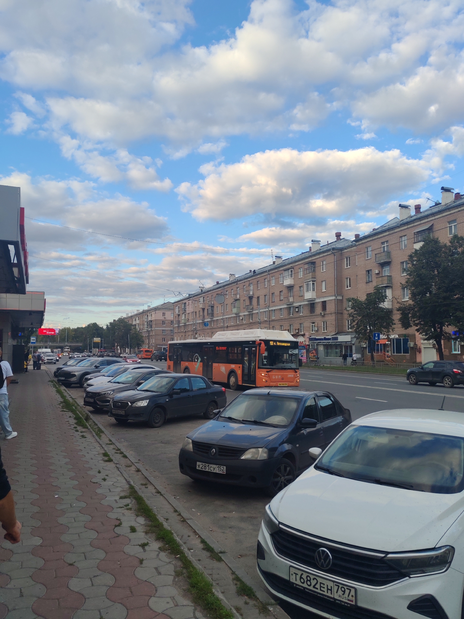 The image shows a street scene in Nizhny Novgorod, Russia. In the foreground, a white Volkswagen car is parked on the right side of the street. To the left of the Volkswagen, a dark gray sedan is parked. Further down the street, several other cars are parked along the sidewalk. An orange bus with the number 93 visible on its front is driving in the street. On the left side of the image, there is a building with a red and white sign. The street is lined with buildings, and there are trees and power lines visible. The sky is blue with white clouds.