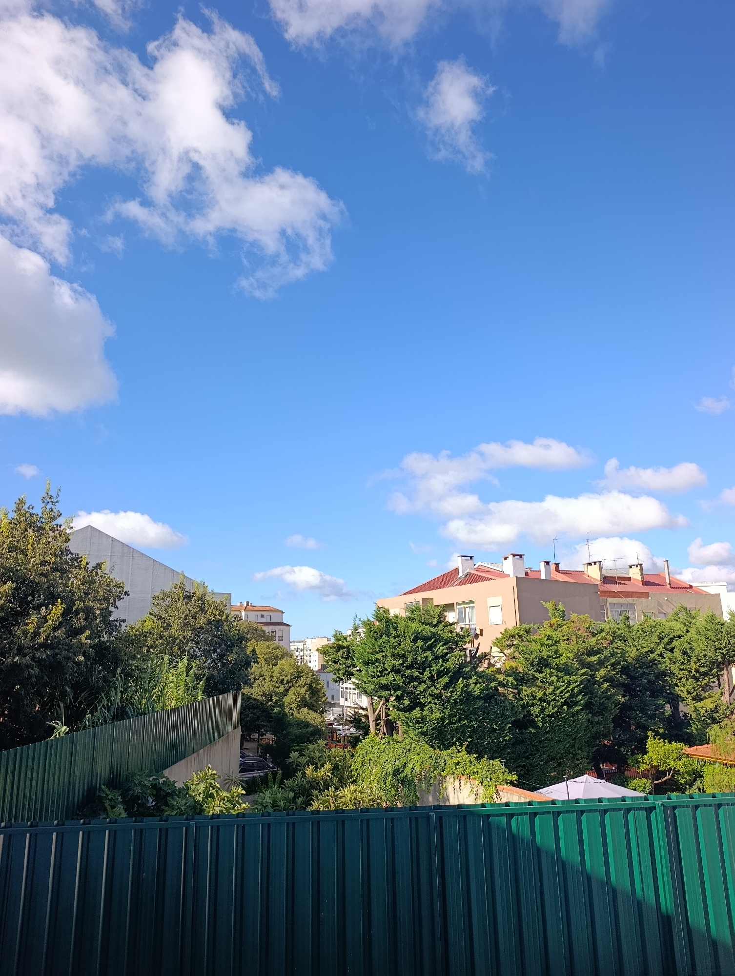 The image depicts a view of urban buildings and trees against a bright blue sky with scattered white clouds. In the foreground, a dark green corrugated metal fence stretches across the bottom of the frame. Behind the fence, more greenery is visible, including lush trees and some buildings peeking through. The buildings appear to be residential or commercial, with red-tiled roofs and light-colored facades. The scene suggests a sunny day in a city. The image does not contain any people or identifiable actions.