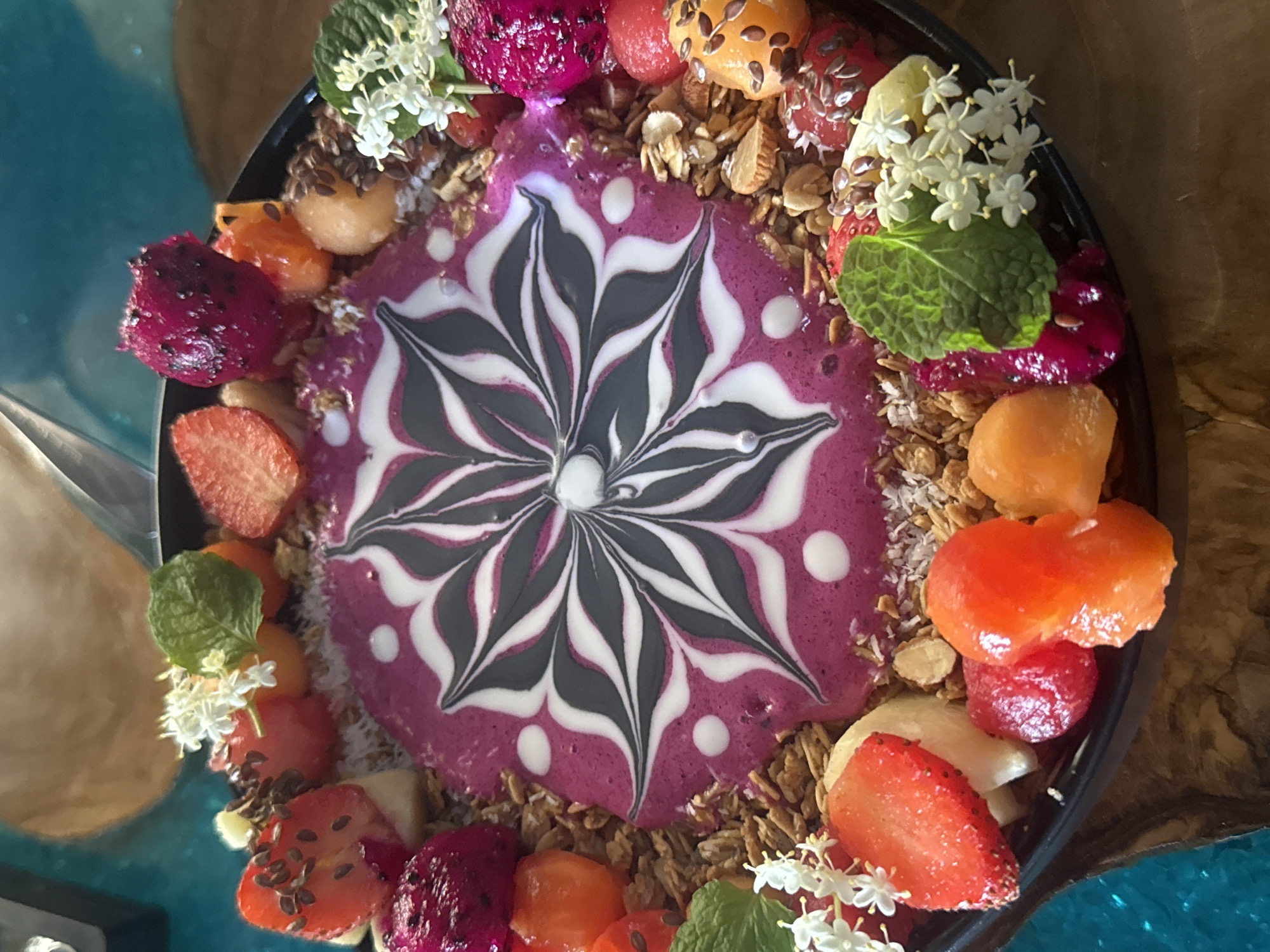 A vibrantly colored smoothie bowl is presented in a black bowl, sitting on a wooden table, potentially in a cafe or restaurant in the Ubud District, Indonesia. The smoothie itself is a bright pink or purple hue, with a striking pattern of white and dark gray swirls resembling a flower or mandala design at the center. Surrounding the smoothie are various fresh fruits, including cut strawberries, pieces of dragon fruit, cantaloupe, and other unidentified fruits. There's also granola scattered around the edges, along with fresh mint leaves and small white flowers used as garnish. The scene evokes a sense of healthy eating and artistic food presentation, in a setting that reflects Ubud's artistic and natural aesthetic.