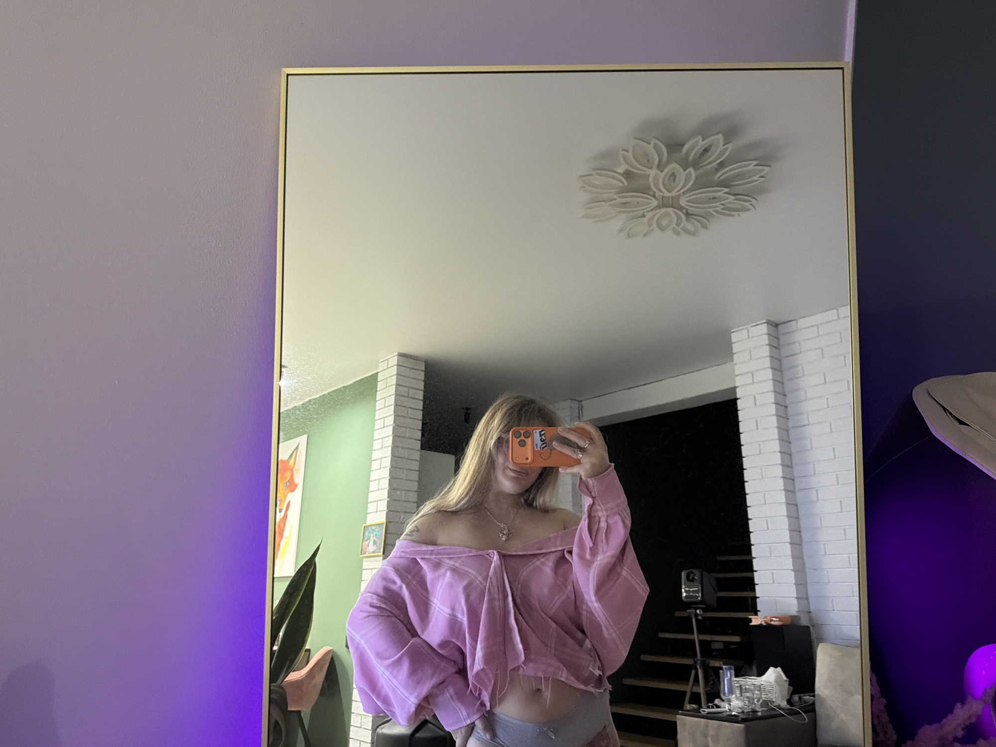 A mirror selfie is captured in what appears to be a residential interior, possibly in Большая Шильна, Russia. The mirror has a gold frame. The background reveals elements such as white brick columns, a staircase, a ceiling light fixture, and a plant. A woman stands in front of the mirror, holding a smartphone in her right hand, partially covering her face with it. She is wearing an off-the-shoulder, oversized pink shirt and grey underwear. The walls are painted in shades of purple and blue, with some walls painted green. There is art visible.
