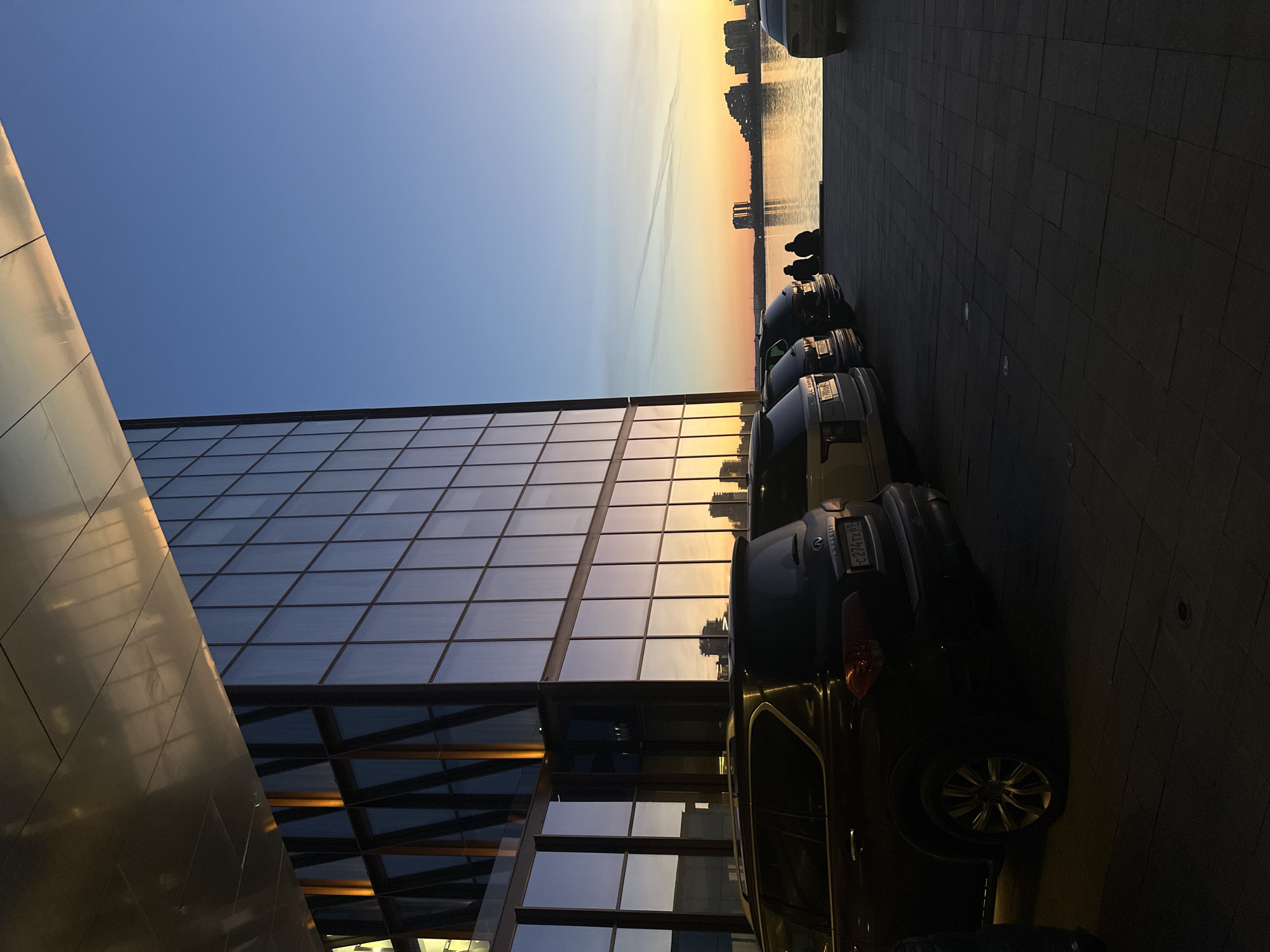 The image depicts an urban waterfront scene at either sunrise or sunset, characterized by a vibrant sky transitioning from blue at the top to warm orange and yellow hues near the horizon, with subtle contrails visible.
On the left, a modern building with a façade composed of large rectangular glass windows and reflective panels dominates the foreground. These surfaces reflect the sky's colors, showing a blend of blue and golden light. Another part of the building features a metallic, angular structure, also reflecting the warm light.
Along the right side of the building, several cars are parked on a dark paved surface. One car, an Infiniti SUV, is clearly visible with its rear facing the viewer. Its license plate displays "C274TX 162," with "162" being the regional code for the Republic of Tatarstan in Russia, where Kazan is located.
Beyond the parked cars and across a body of water, a city skyline composed of various buildings is silhouetted against the colorful sky. Two human figures are visible in silhouette by the water's edge, appearing to be seated or standing and observing the vista. The ground in the foreground on the right is paved with dark, rectangular blocks. The overall lighting casts long shadows, enhancing the reflective qualities of the building and water, creating a serene and picturesque twilight atmosphere.
