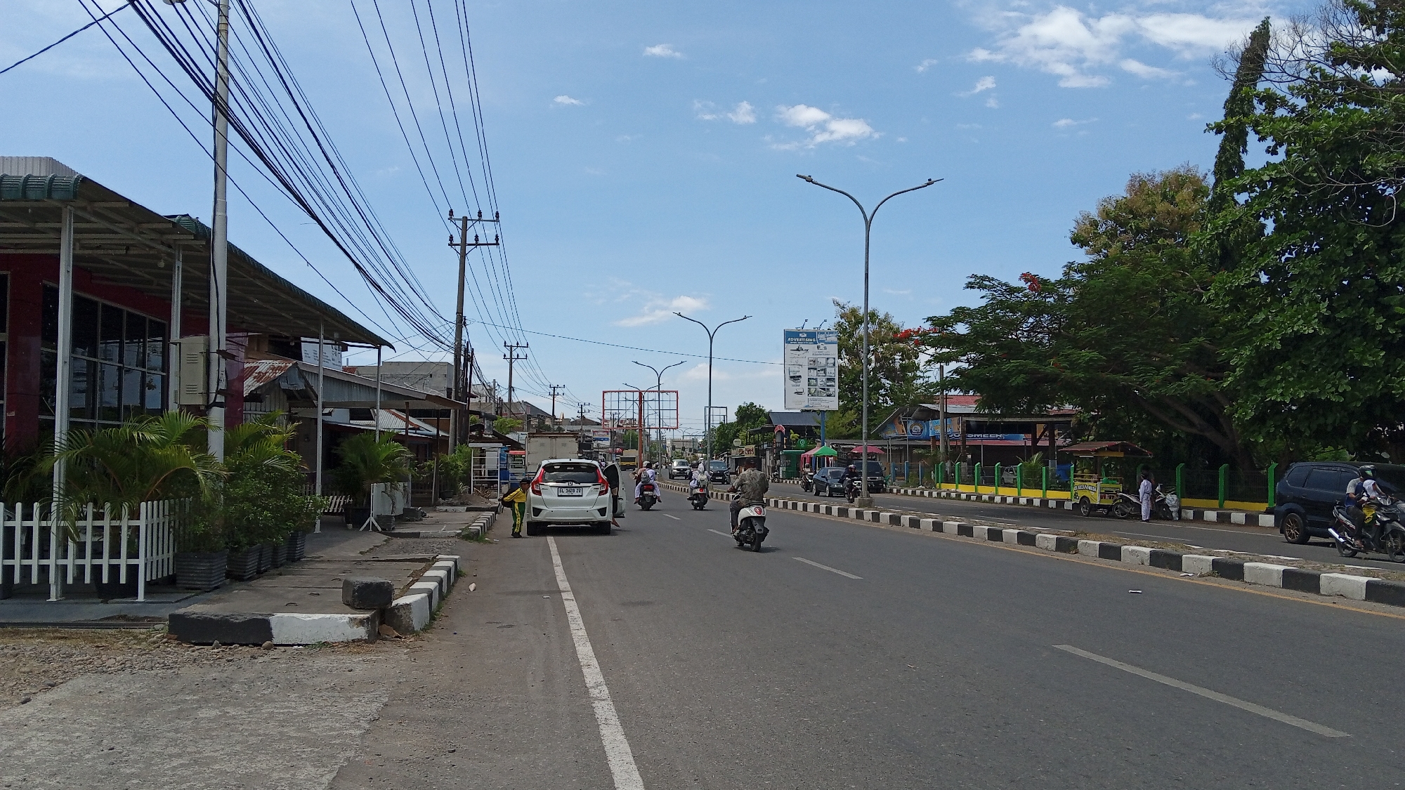 The image depicts a street scene in Julikeude Dua, Indonesia, on a sunny day with scattered clouds. A paved road runs horizontally across the frame, with traffic lanes marked by white lines. Several vehicles are present, including a white car, multiple motorcycles, and a black SUV. Some people are seen riding motorcycles, and one person is standing near a white car.
On the left side of the road, there are several commercial buildings with awnings and storefronts. A white fence is visible in front of some potted plants. Electrical poles and numerous overhead wires dominate the upper left portion of the image.
On the right side of the road, there are more buildings, some with colorful awnings, and trees. A fence separates the sidewalk from the road. Streetlights are positioned along the road. The overall environment suggests a busy urban or semi-urban area.