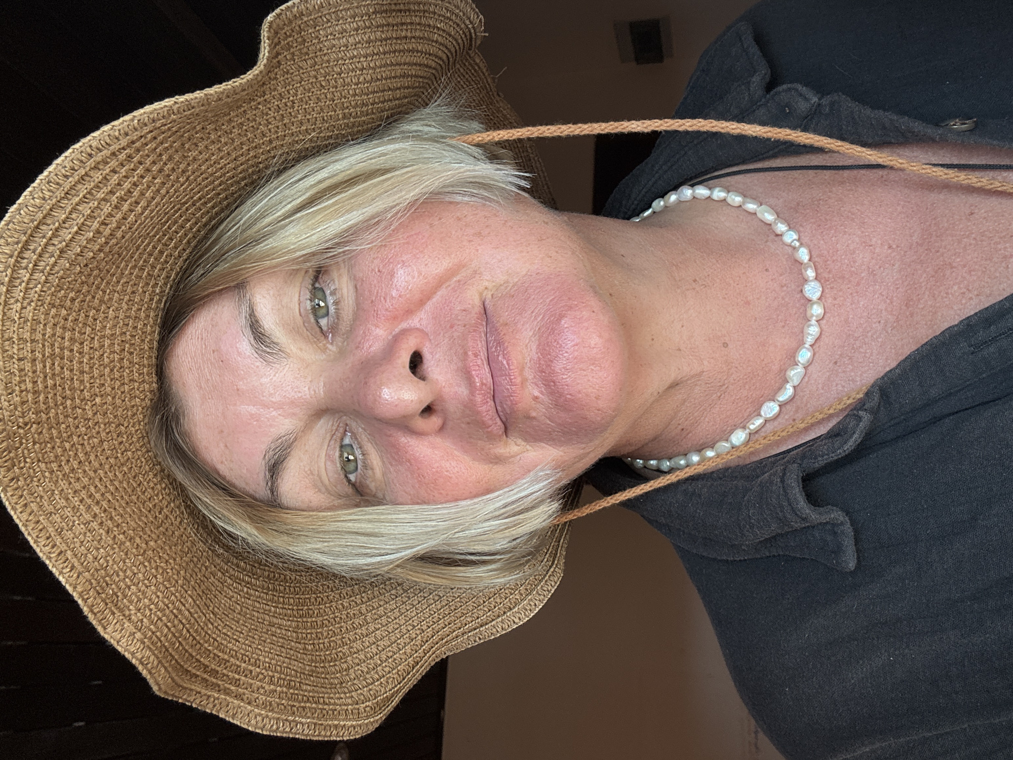A woman with fair skin and short blonde hair is wearing a brown woven sun hat and a strand of pearls. She is wearing a dark-colored shirt. The image is a close-up portrait, possibly a selfie, with the woman angled slightly to the side. The background is out of focus. There is no indication of the location being บ้านบางรักษ์, Thailand, or any other specific location.