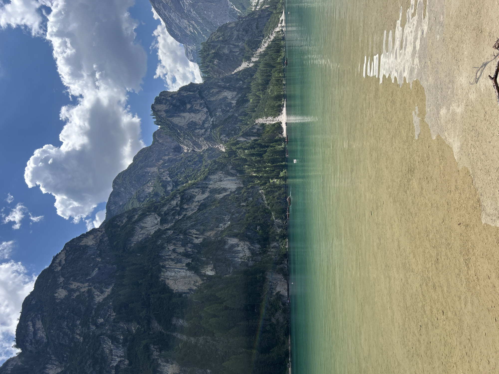 The image depicts a scenic view of a lake with a mountain backdrop. The lake's water is a clear, greenish-blue hue, with a sandy shore visible on the right side of the frame. Sunlight reflects off the water's surface, creating shimmering patterns. In the distance, a rugged, imposing mountain range rises, partially covered in lush green trees. The sky above is a vibrant blue, dotted with fluffy white clouds. A few small boats are scattered across the water, suggesting a leisurely activity. The overall atmosphere is serene and tranquil, evoking a sense of natural beauty and peacefulness. The time of day appears to be midday, given the bright sunlight. No visible text is present in the image.