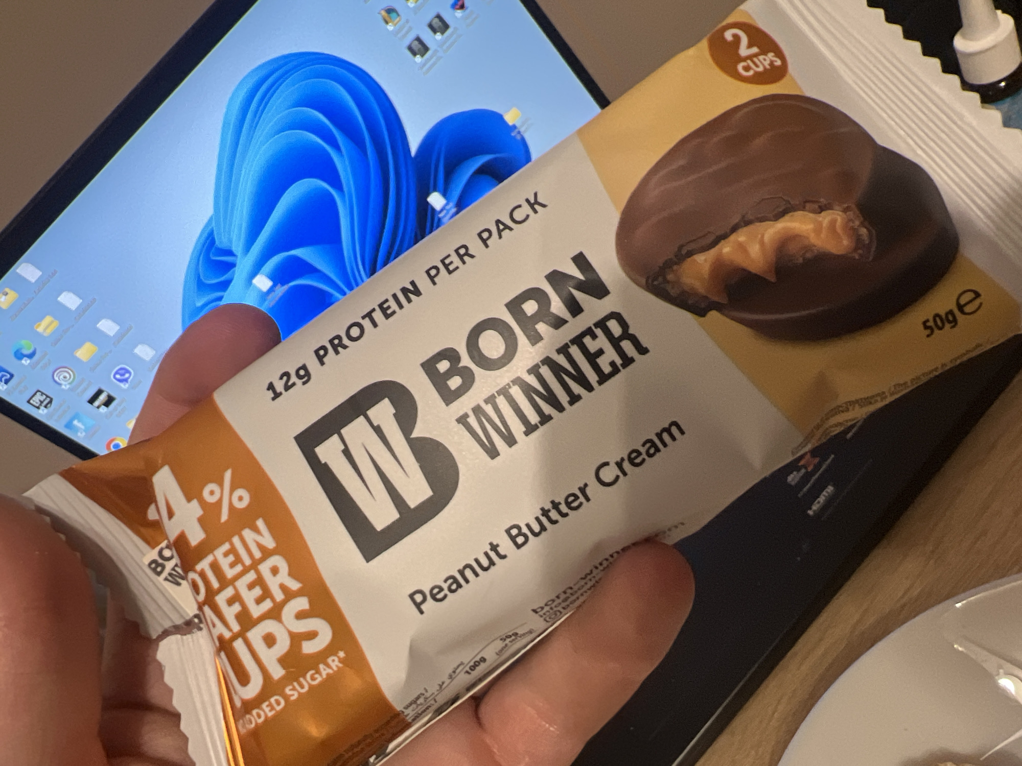 A close-up, high-angle indoor shot depicts a person's hand holding a "Born Winner" brand protein snack package. The package is white and orange, displaying the text "PROTEIN WAFER CUPS," "Peanut Butter Cream," "12g PROTEIN PER PACK," "4% ADDED SUGAR*," and "50g e." An illustration on the package shows two chocolate-coated peanut butter cups, one revealing its creamy peanut butter interior, accompanied by "2 CUPS" in an orange oval. The background reveals a laptop screen displaying the Windows 11 desktop with a vibrant blue, abstract, swirling wallpaper. Multiple application icons, including Google Chrome, Epic Games Launcher, and a purple chat application icon, are visible on the desktop. The laptop's dark bezel and a faint "HDMI" label on its side are also present. A light wooden surface serves as a desk or table, partially obscured by the laptop and a white ceramic plate with a metallic utensil on its left side. The specific location within Sofia, Bulgaria, cannot be identified from the visual evidence; however, the "Born Winner" brand and the potential presence of the Viber application icon (the purple chat icon) suggest a Bulgarian context.
